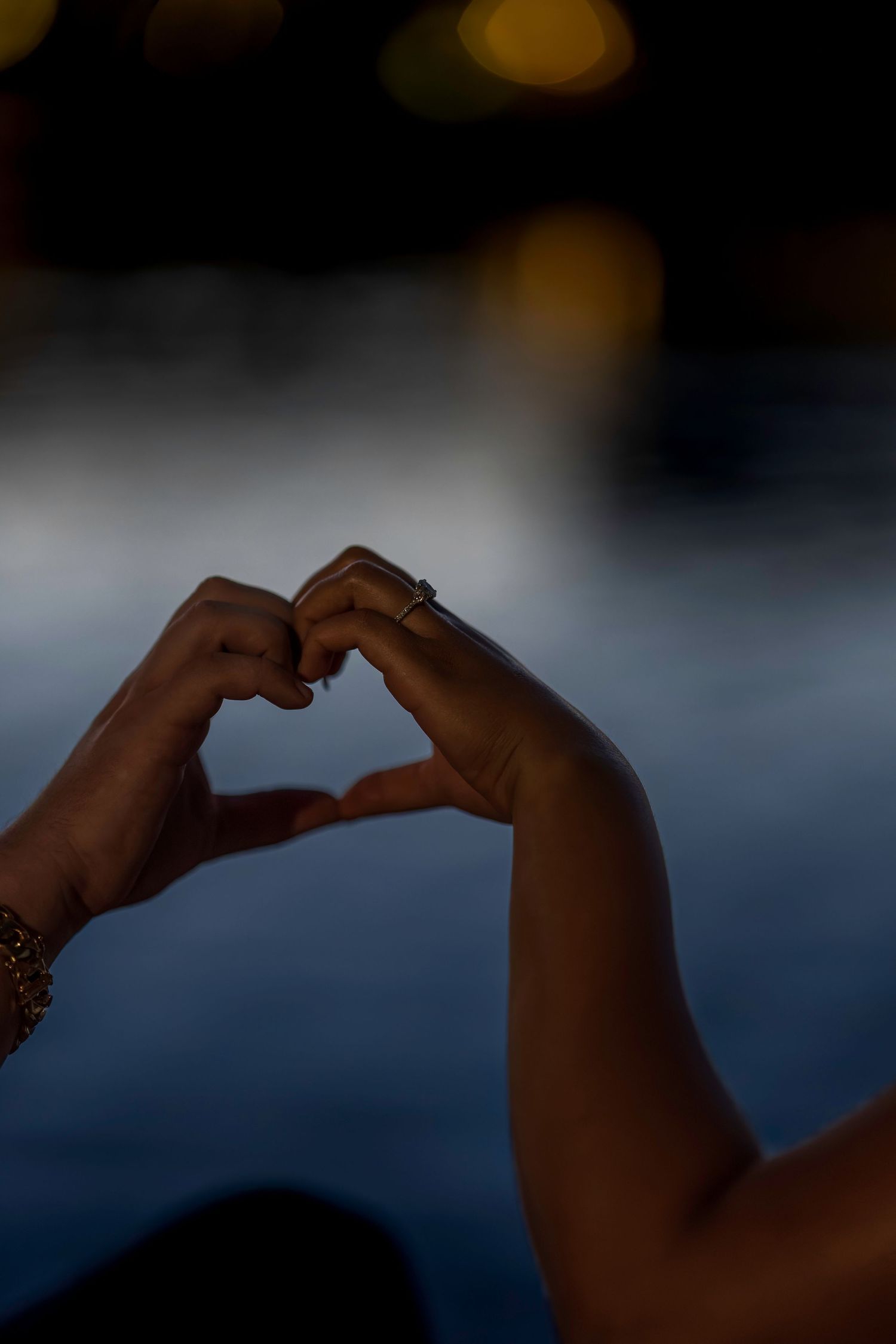 Two hands forming a heart shape at dusk with bokeh lights in the background, one hand wearing a ring.