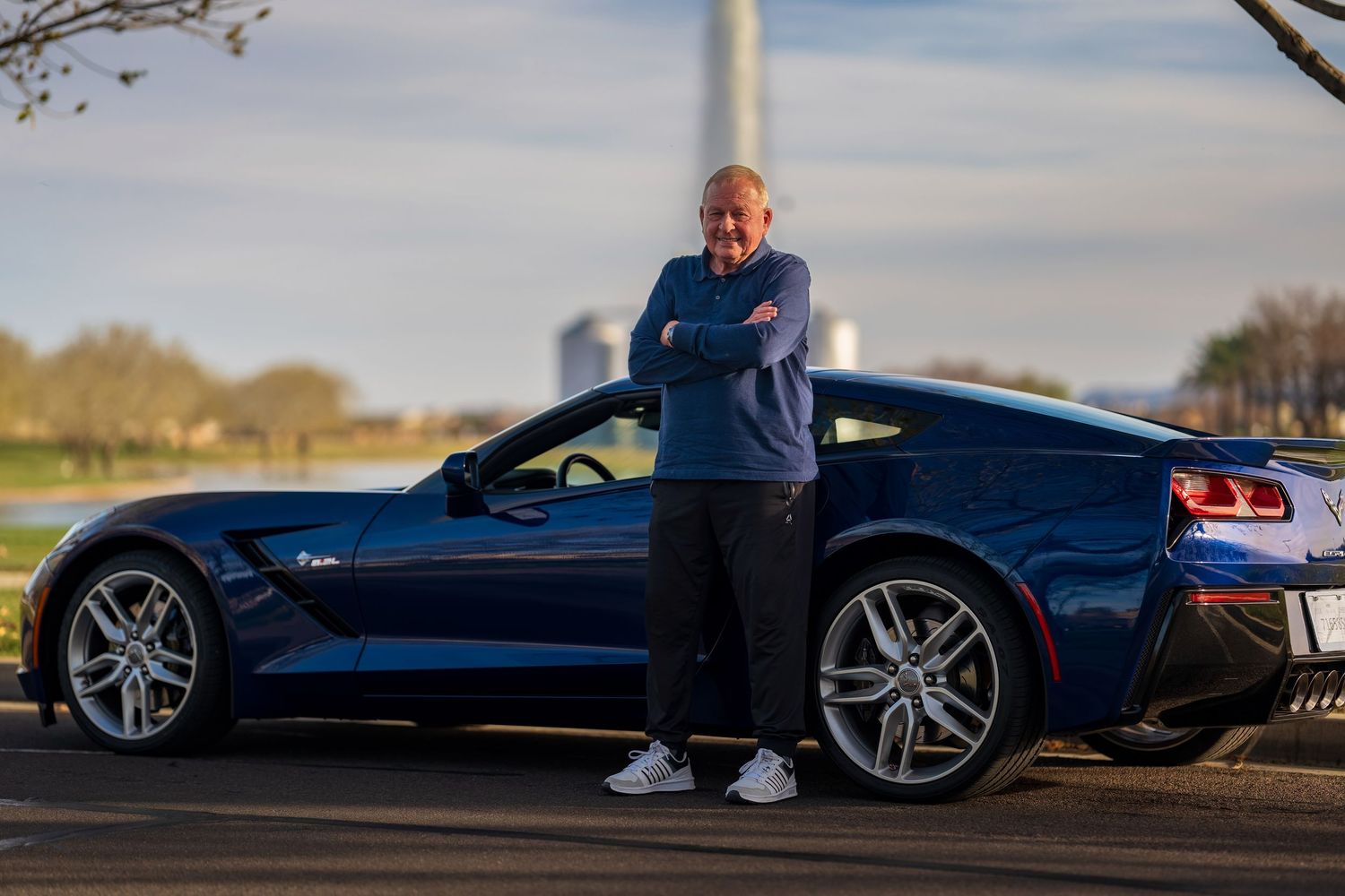 Older man in blue sweater stands with arms crossed beside a navy Chevrolet Corvette Stingray outdoors.