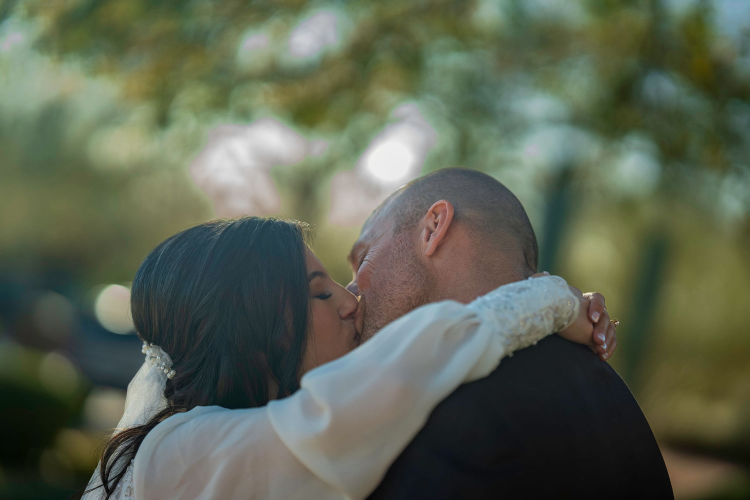 Newlywed couple shares romantic kiss outdoors surrounded by soft bokeh background.