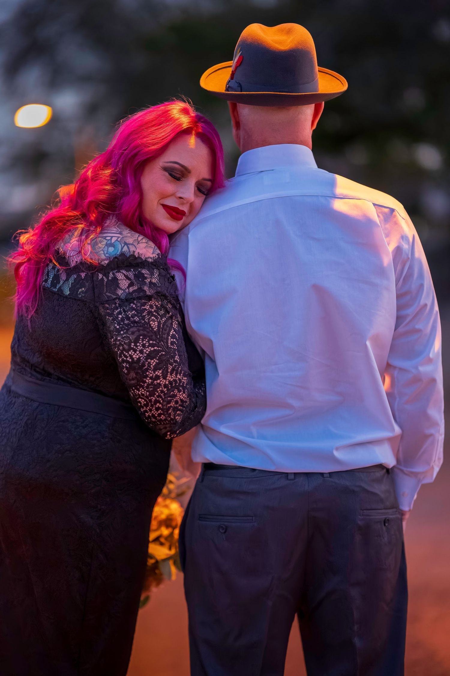 A woman with pink hair in a black lace dress leans on a man in a white shirt and gray fedora at dusk.