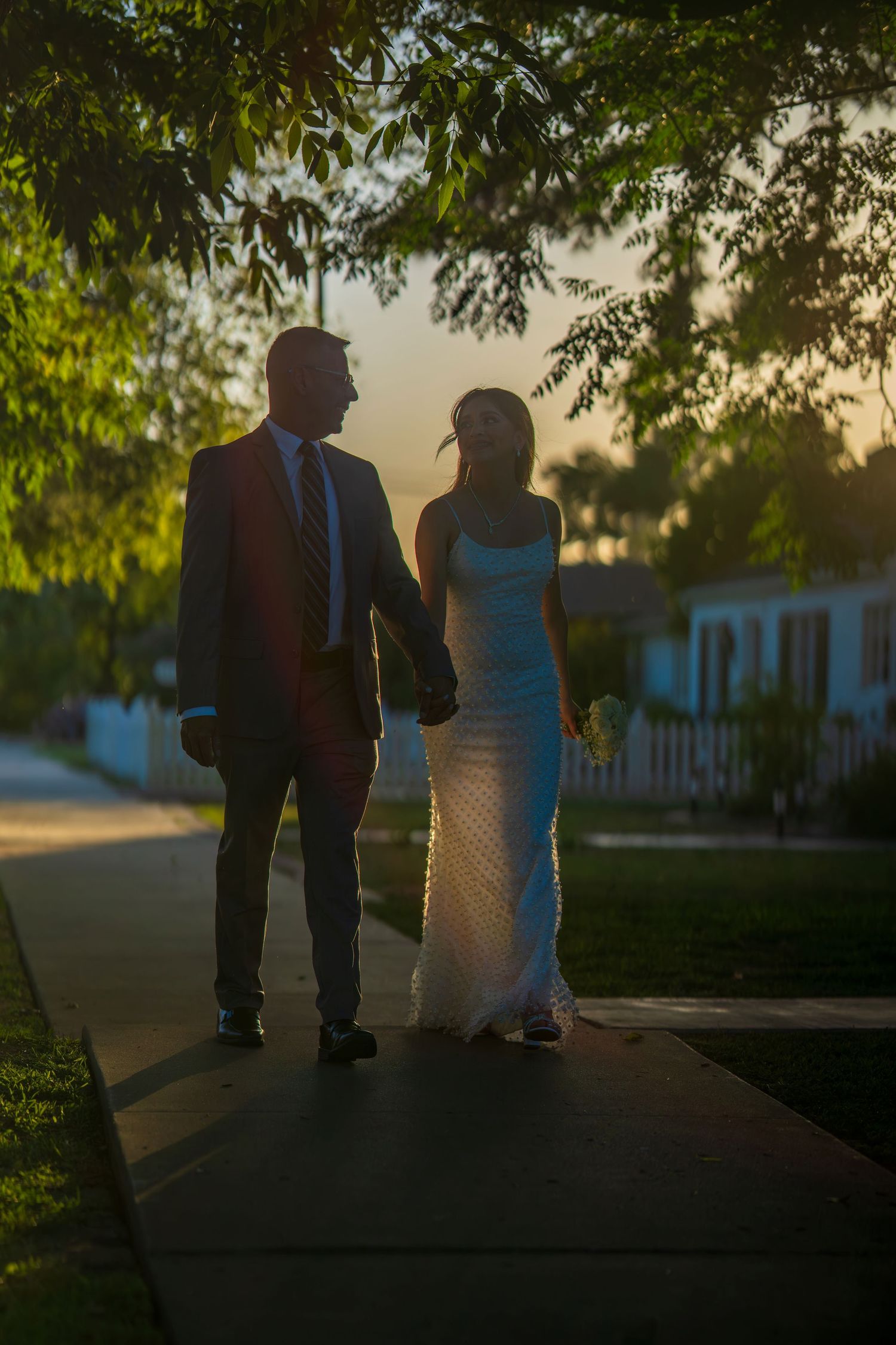 Bride and groom hold hands walking at sunset under green trees in a romantic silhouette.