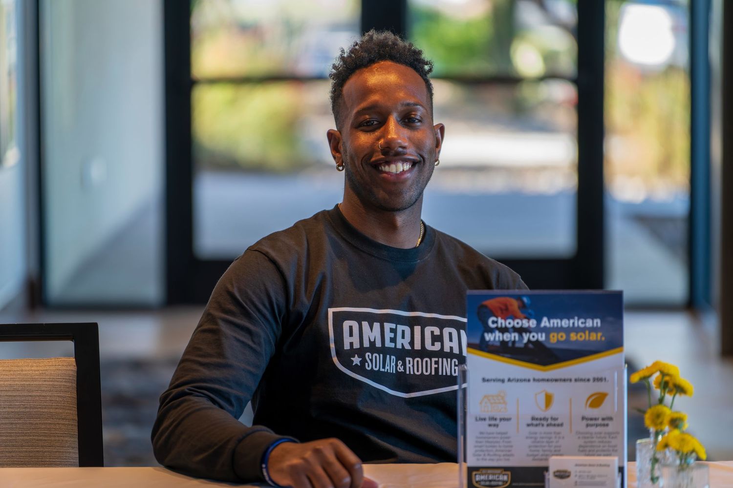 Professional in black American Solar Roofing shirt sitting at desk with company materials.