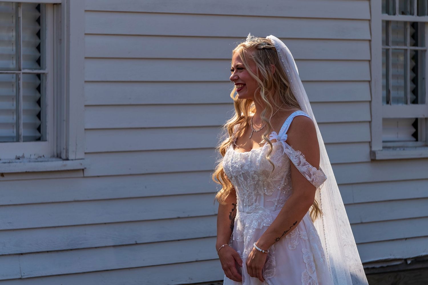 Bride in lace wedding gown and veil walking outside a white church building.