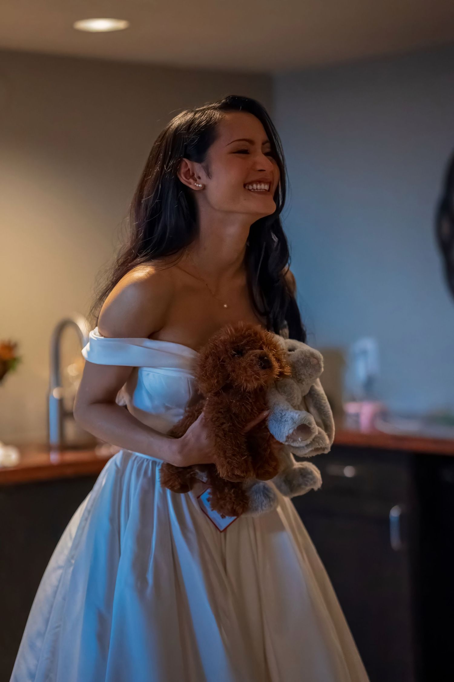 A bride in an off-shoulder wedding dress holds a teddy bear while smiling in an indoor setting.