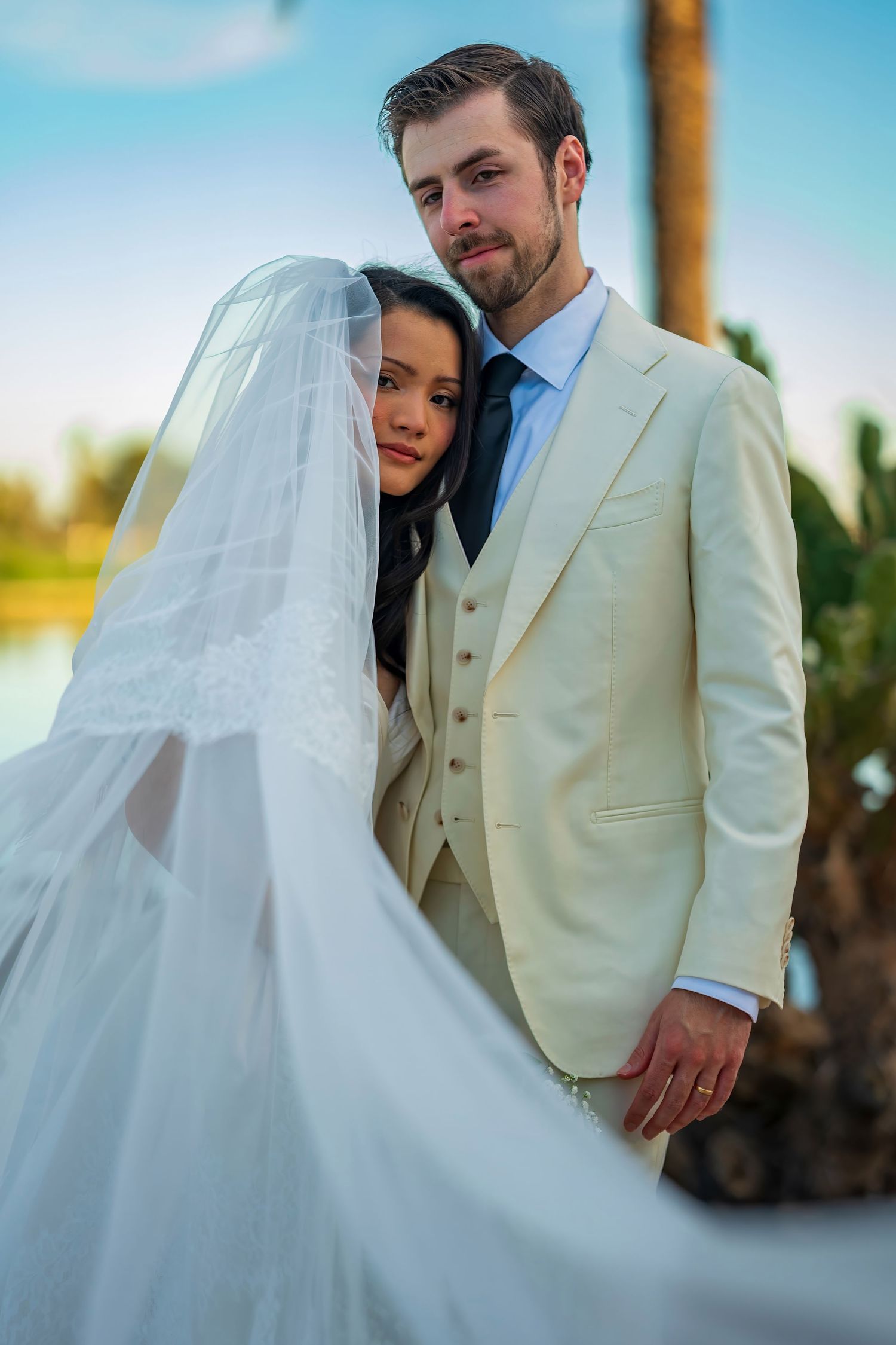 Romantic wedding couple embraces outdoors with flowing veil against blue sky.