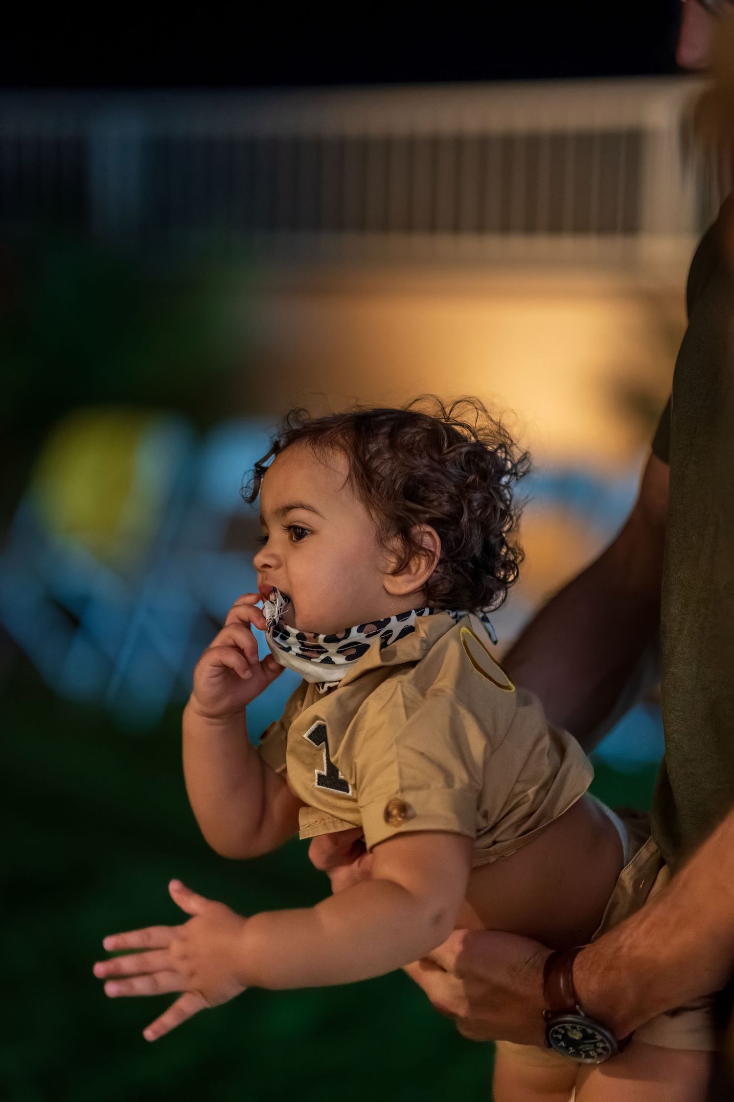 A small child in casual wear putting fingers in their mouth while being held.