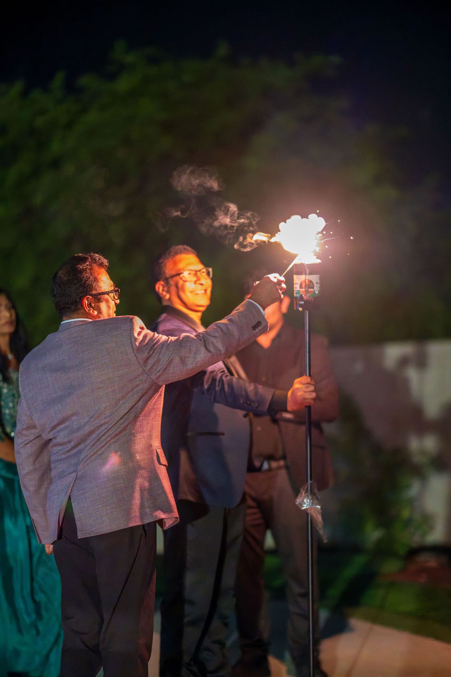 Two people in suits light sparklers together during a nighttime celebration.