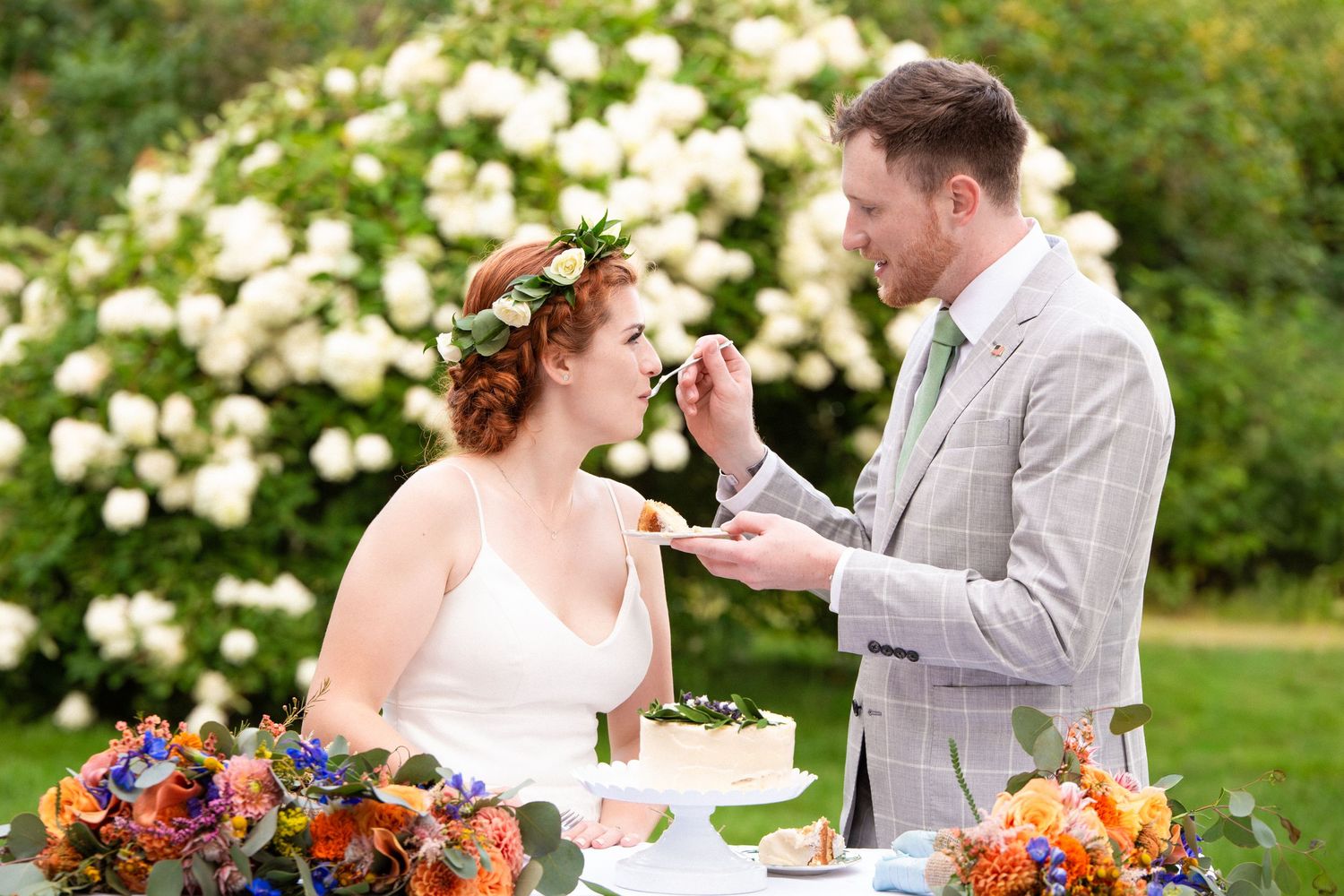 Groom feeds cake to redhead bride with flower crown at outdoor garden wedding reception, Xenia Helix Photography, Boston, MA