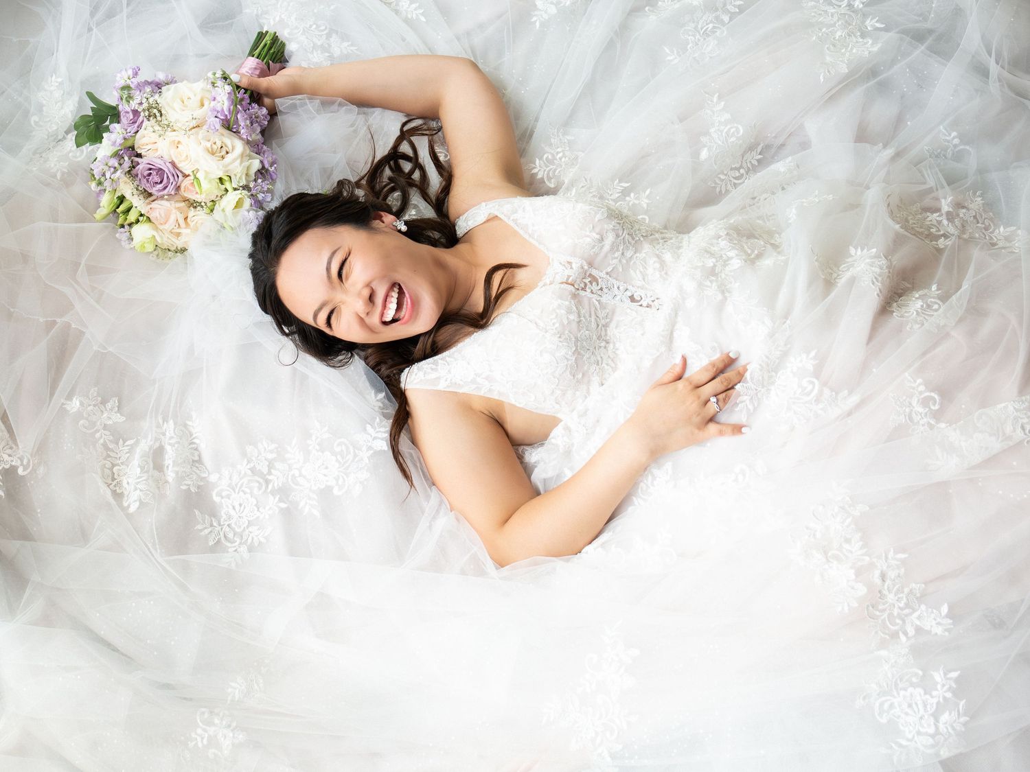 Joyful bride in lace ball gown lies in dress holding purple and white bouquet laughing, The Cape Club of Sharon, Sharon, MA