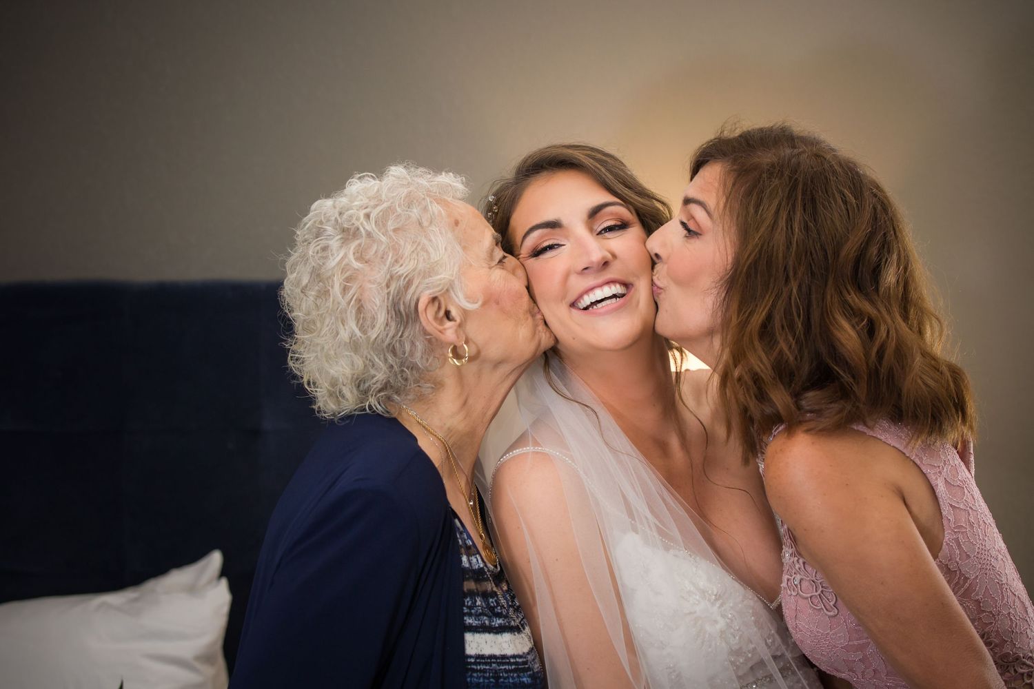 A smiling bride receives kisses on both cheeks from her grandmother and mother before wedding, Hotel 1620 Plymouth Harbor, Plymouth, MA