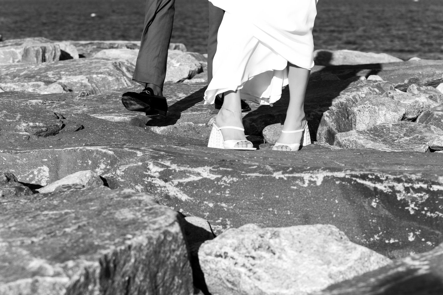 Black and white close-up of bride and groom's feet walking on rugged coastal rocks at wedding, Scituate Lighthouse, Scituate, MA