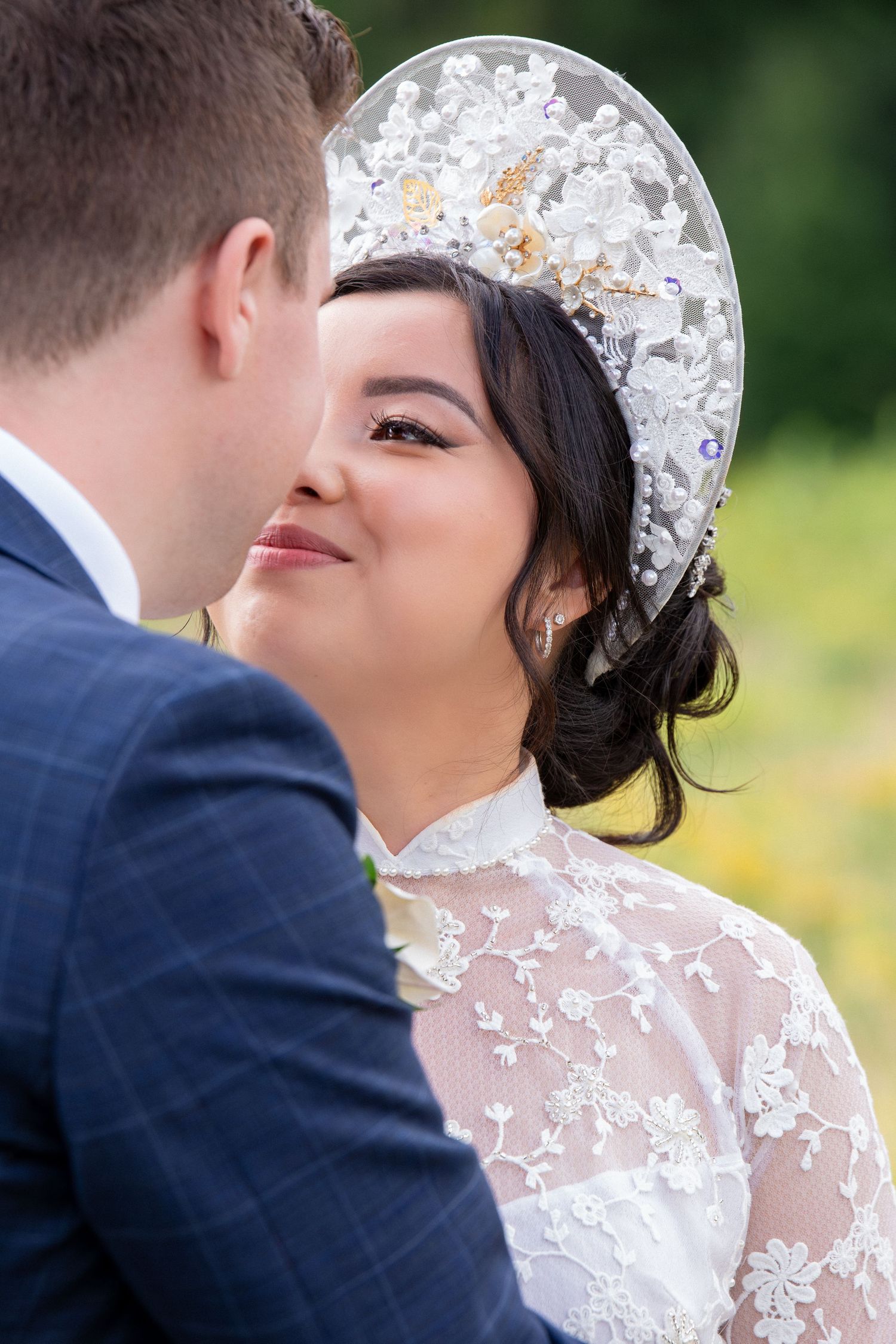 Bride in a white lace dress and a Vietnamese floral headpiece smiling at the groom during the wedding, Harrington Farm, Princeton, MA