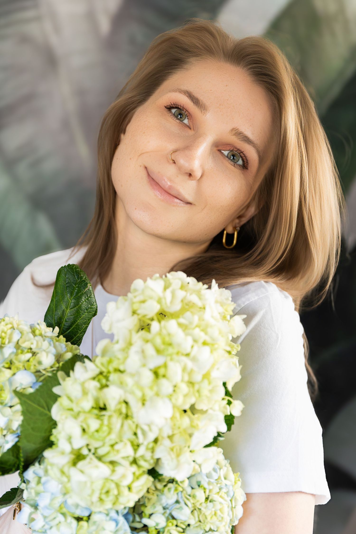 Young blonde woman with green eyes holding a bouquet of white and green hydrangeas, smiling softly at the camera.