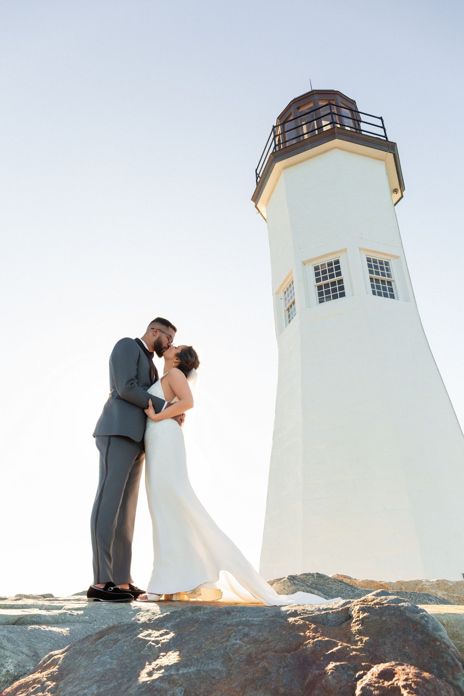 Bride and groom share a romantic kiss in front of a white lighthouse on rocky shores, Scituate Lighthouse, Scituate, MA