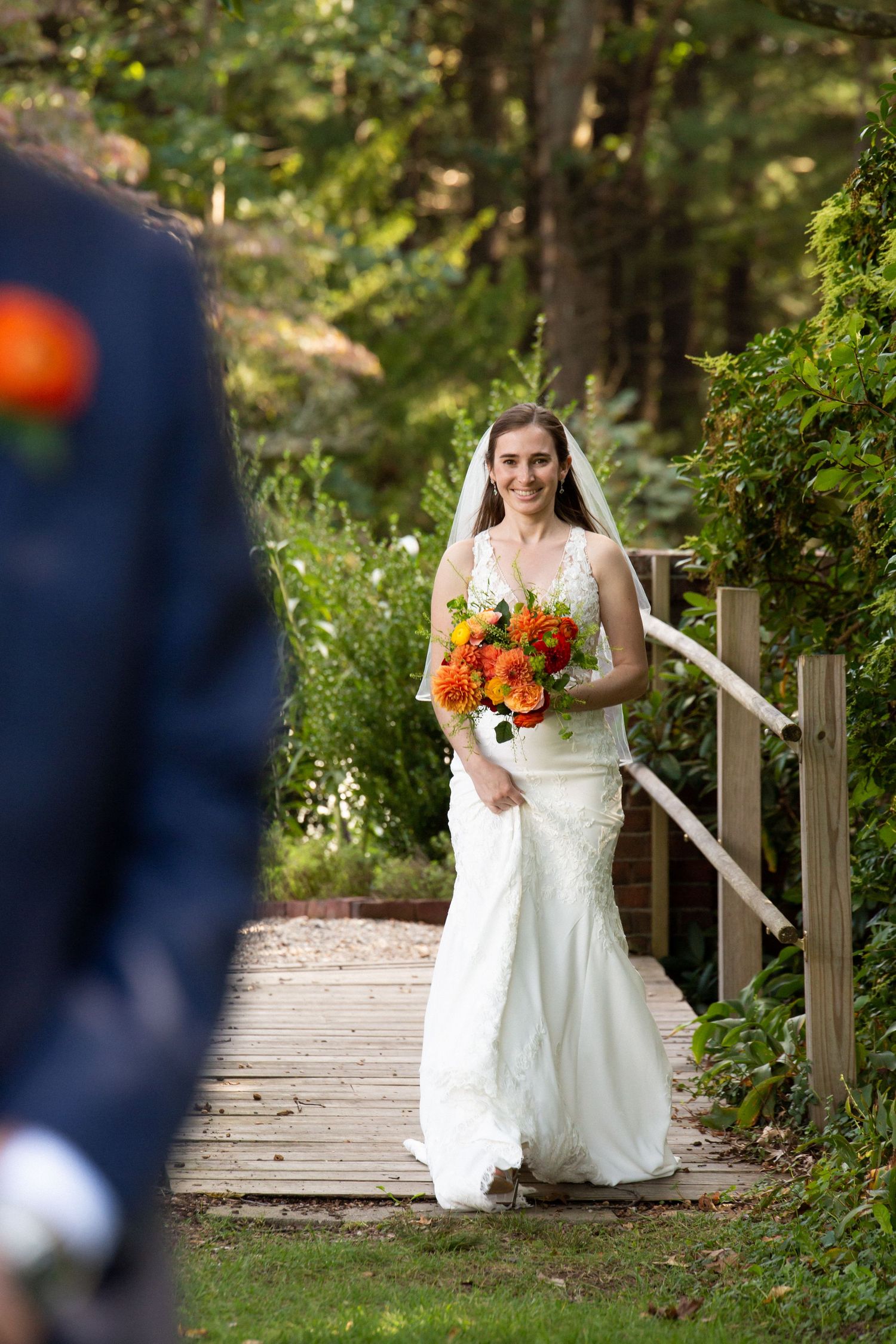 Radiant bride in lace gown carrying orange bouquet walking along a wooden garden path toward groom, The Bradley Estate, Canton, MA