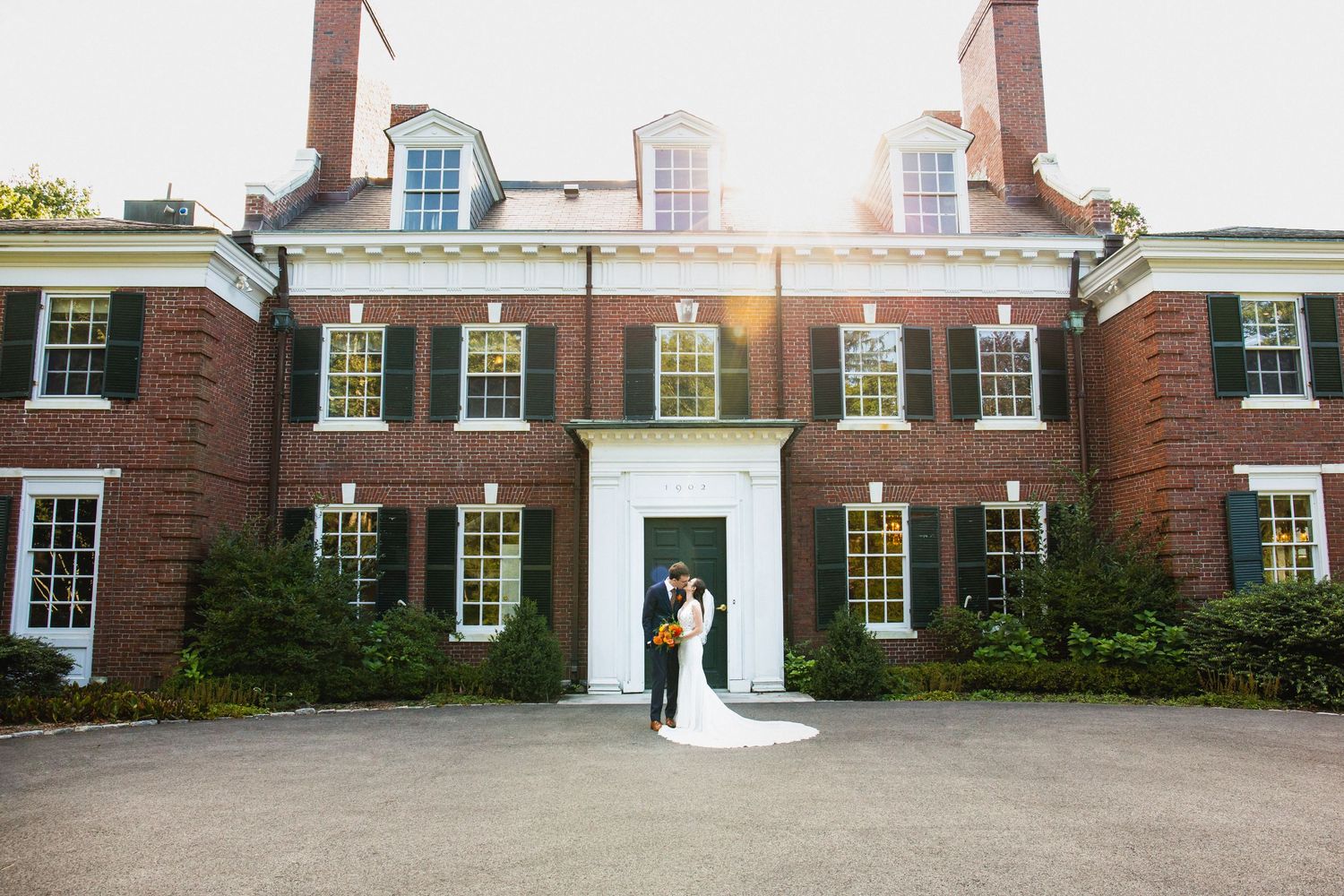 Wedding couple posing in front of red brick colonial mansion with white trim and dark green shutters, The Bradley Estate, Canton, MA