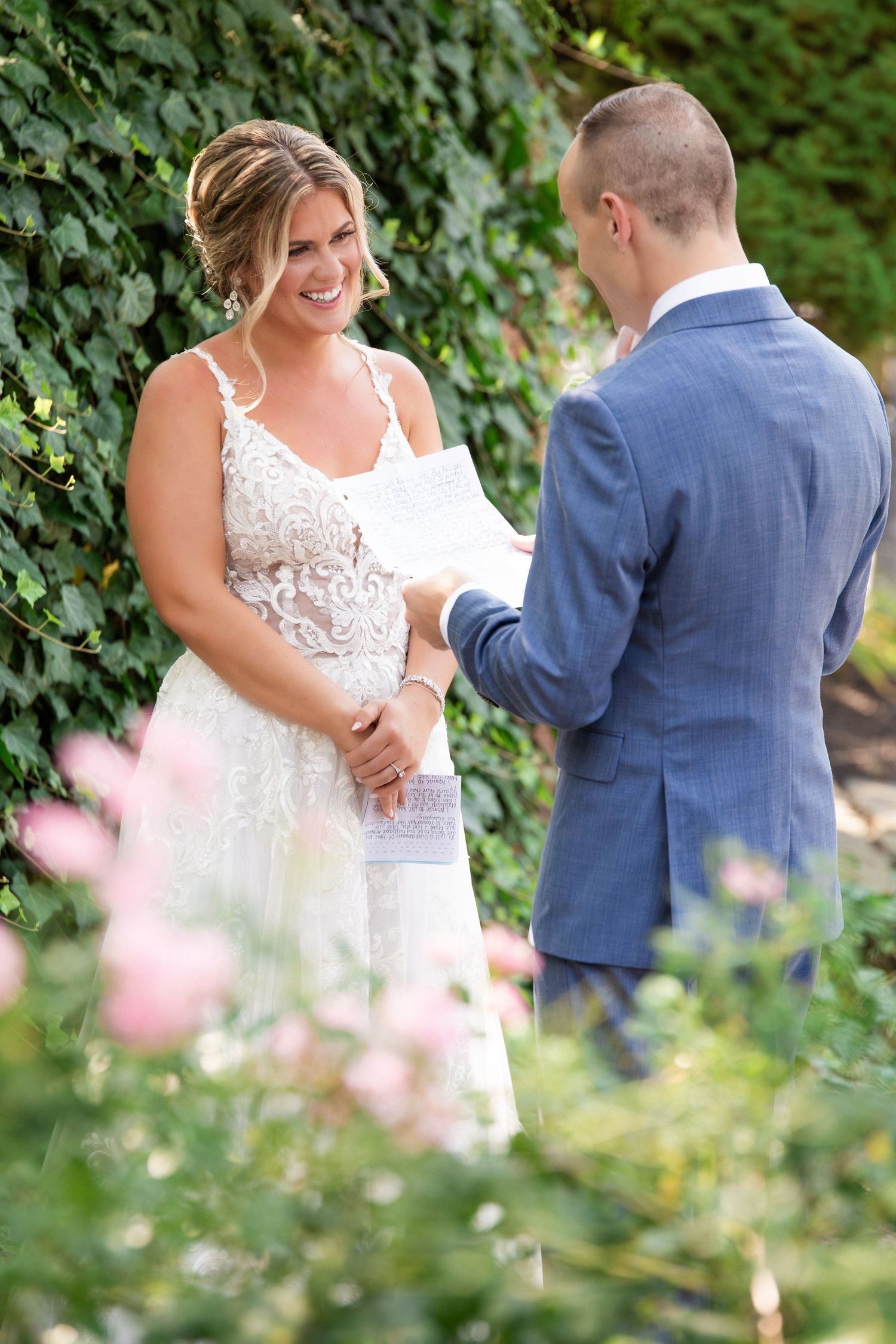 Smiling bride in lace wedding dress reads private vows to groom in blue suit in the garden, The Red Lion Inn, Cohasset, MA