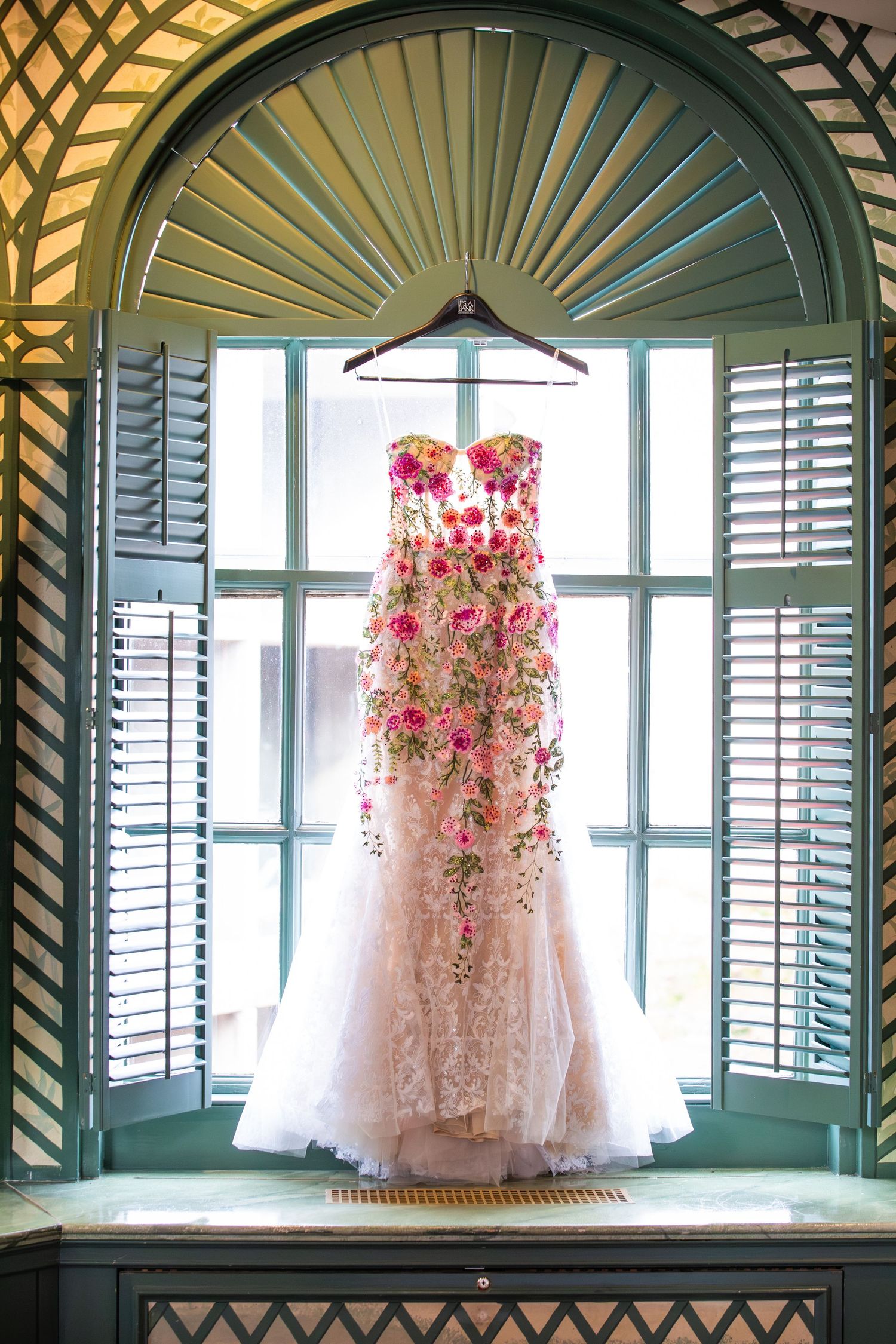 Blush and pink floral-embroidered strapless wedding dress hanging in an arched, green-shuttered window, Harvard Faculty Club, Cambridge, MA