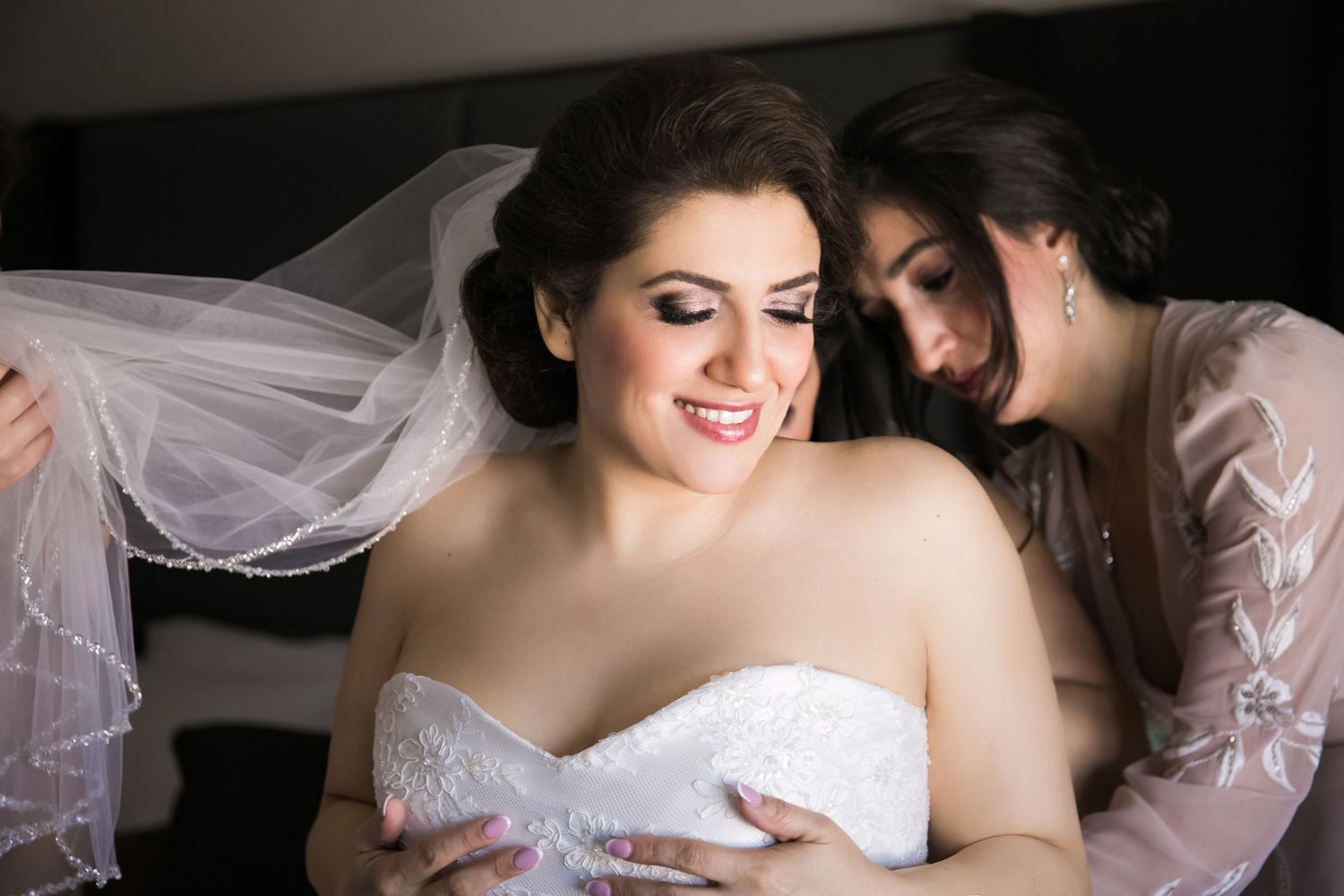 Bride in strapless lace gown smiling as bridesmaid adjusts her veil during wedding prep, Hilton Garden Inn Foxborough Patriot Place, MA
