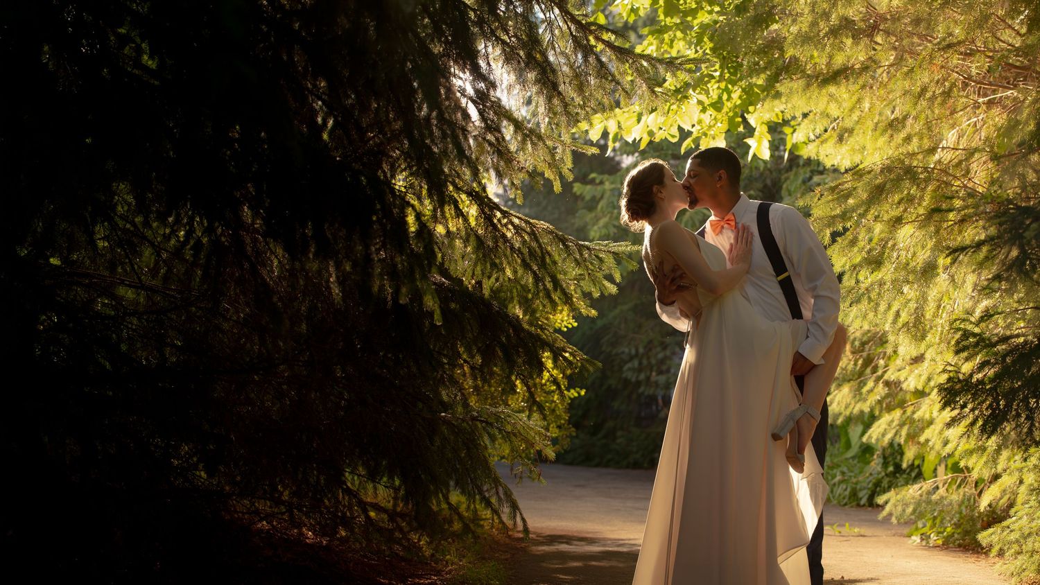 A bride and groom share a romantic kiss surrounded by lush green trees in a sunlit garden setting,  Groveland Fairways, Groveland, MA