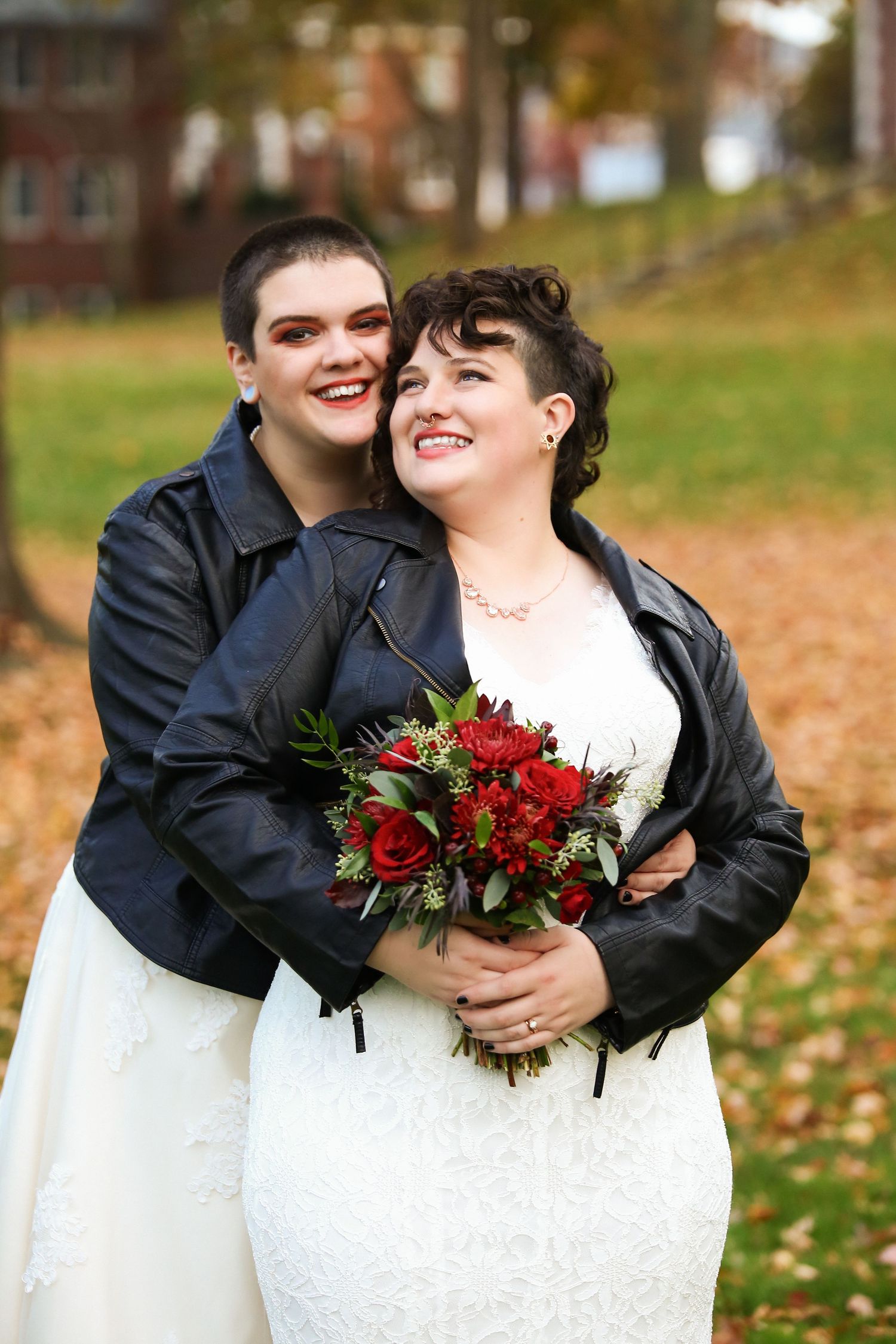 Two brides in white gowns and leather jackets embracing outdoors surrounded by colorful autumn foliage, Inn on Boltwood, Amherst, MA