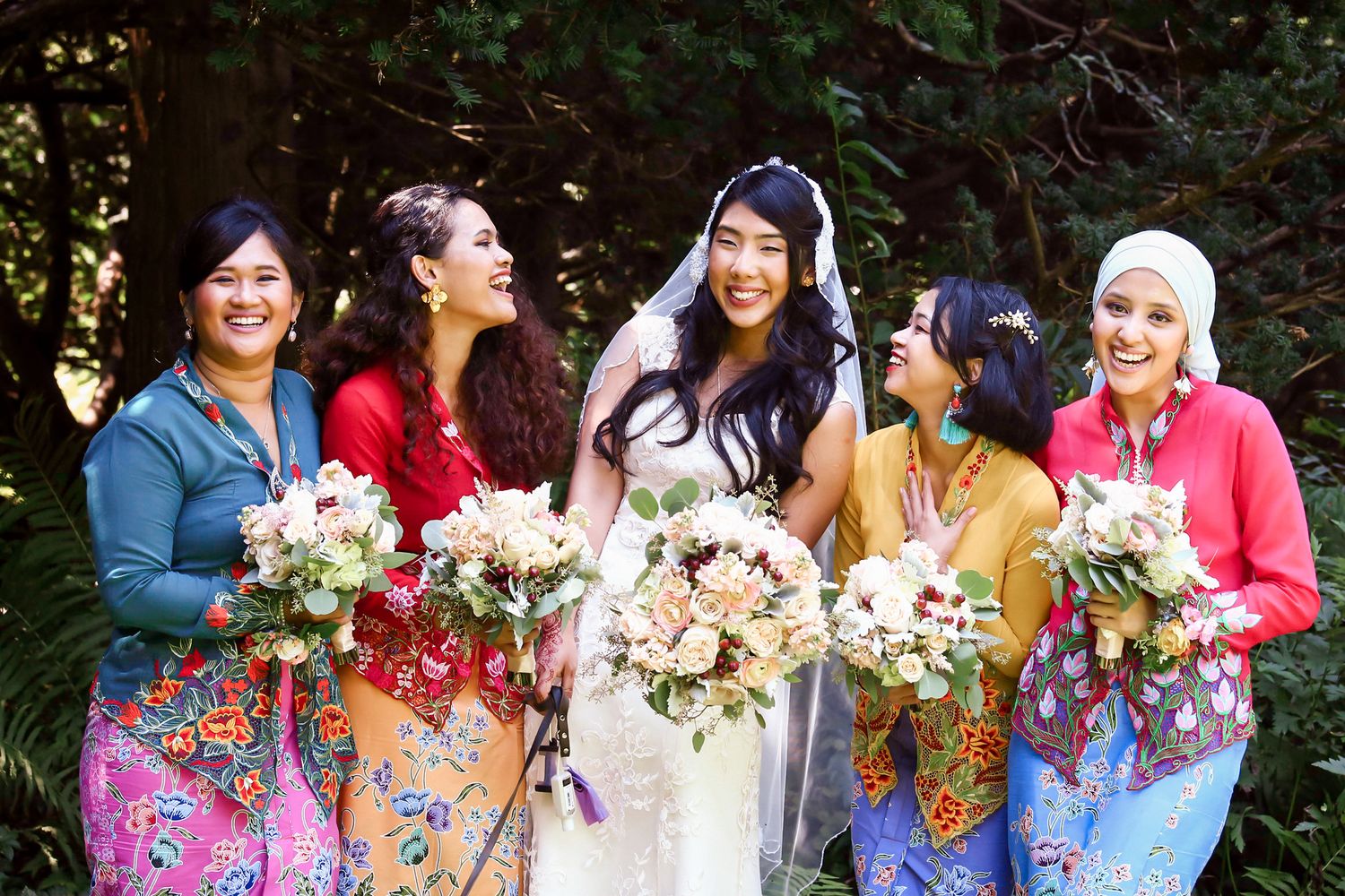 Bride in white gown stands with four bridesmaids in colorful floral kebaya holding bouquets outdoors, Mass Audubon's Habitat, Belmont, MA