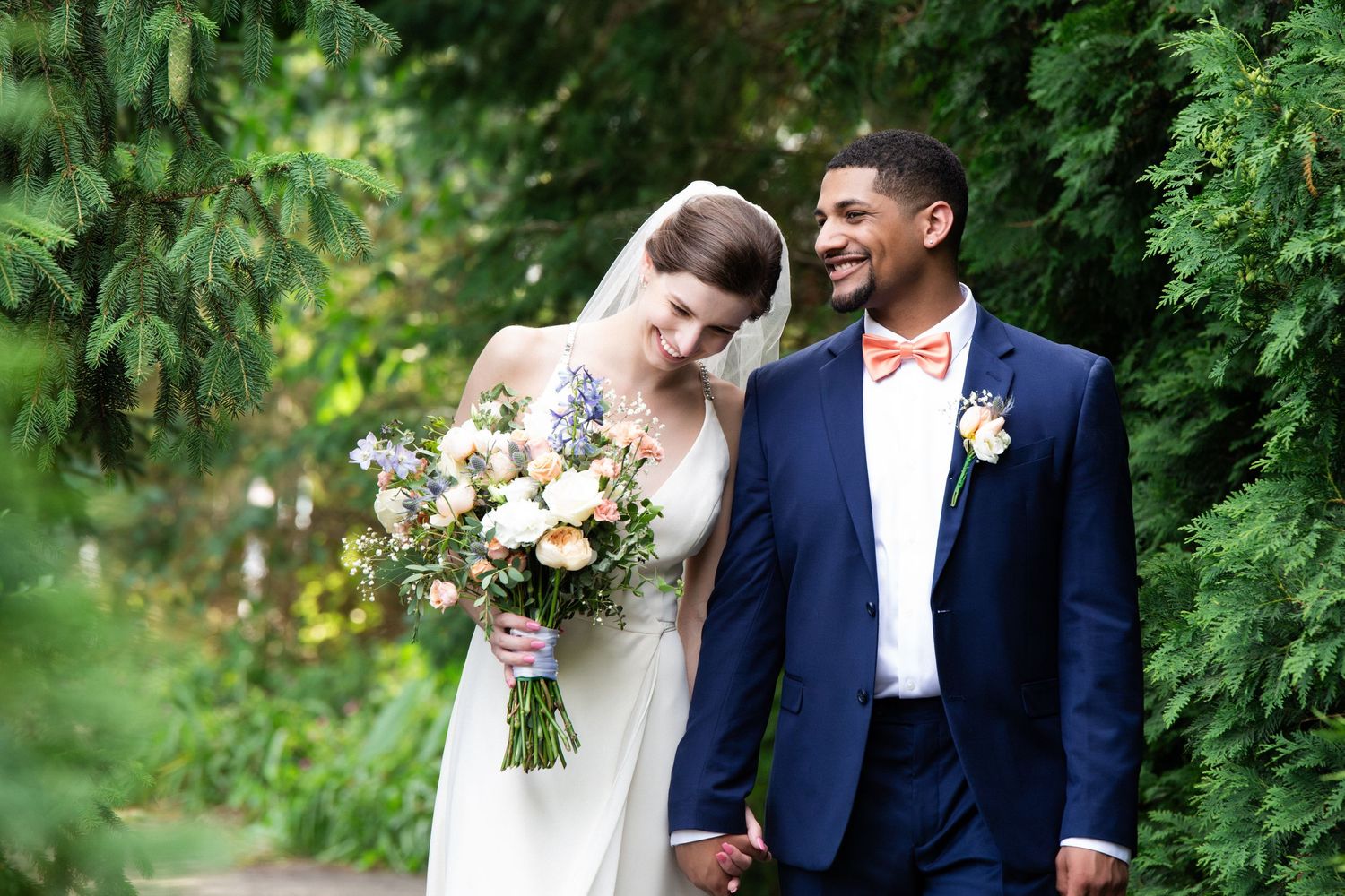 Groom in navy suit with coral bow tie smiles at bride holding a pastel bouquet in the garden, Groveland Fairways, Groveland, MA