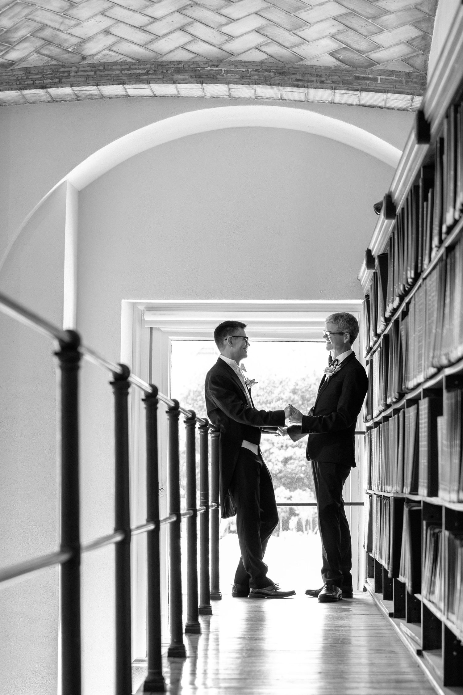 Black and white photo of two grooms in suits holding hands in arched library hallway, Guastavino Room, Boston Public Library, Boston, MA