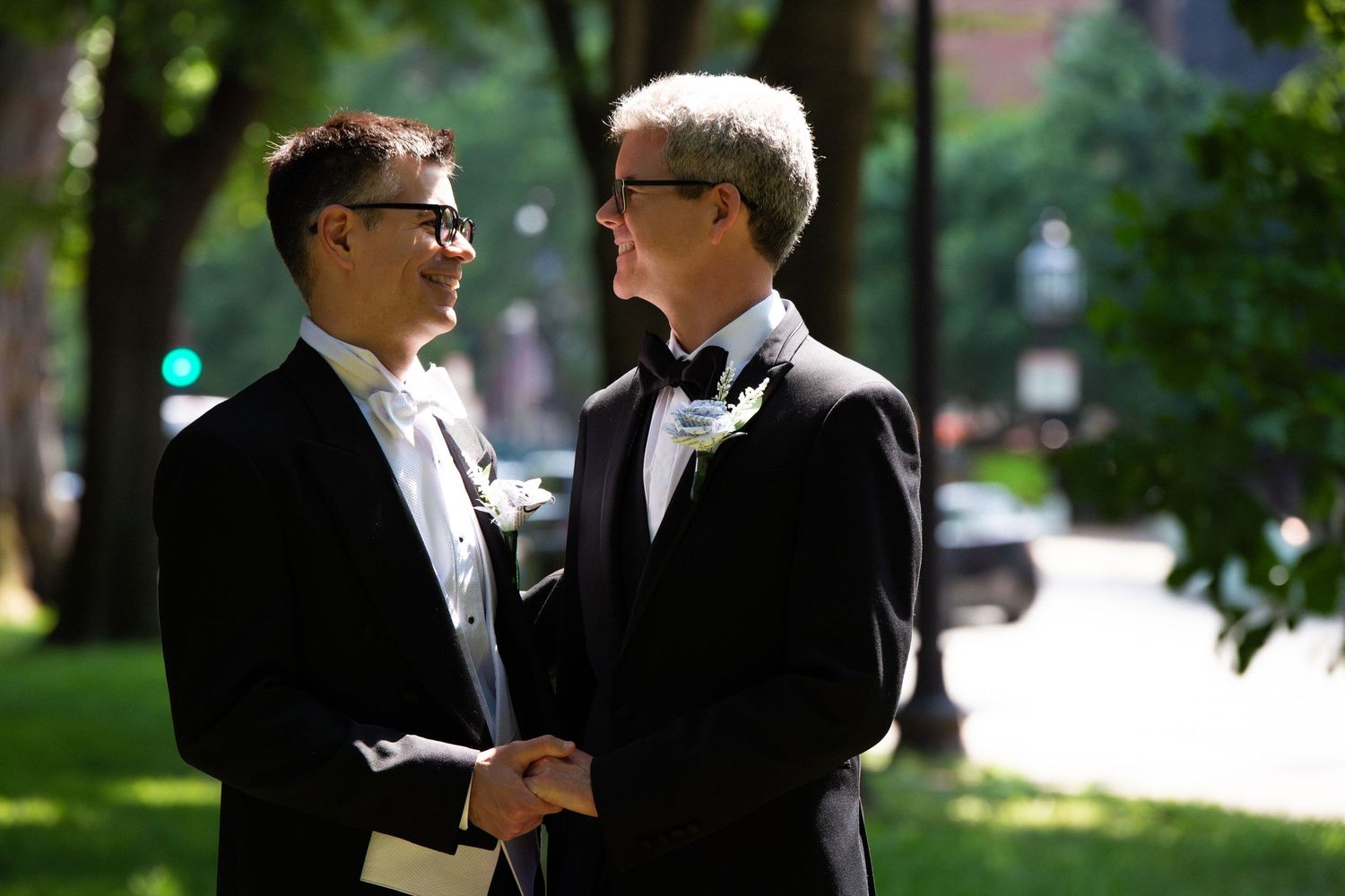 Two grooms in tuxedos holding hands and smiling at each other outdoors on their wedding day, Commonwealth Avenue Mall, Boston, MA