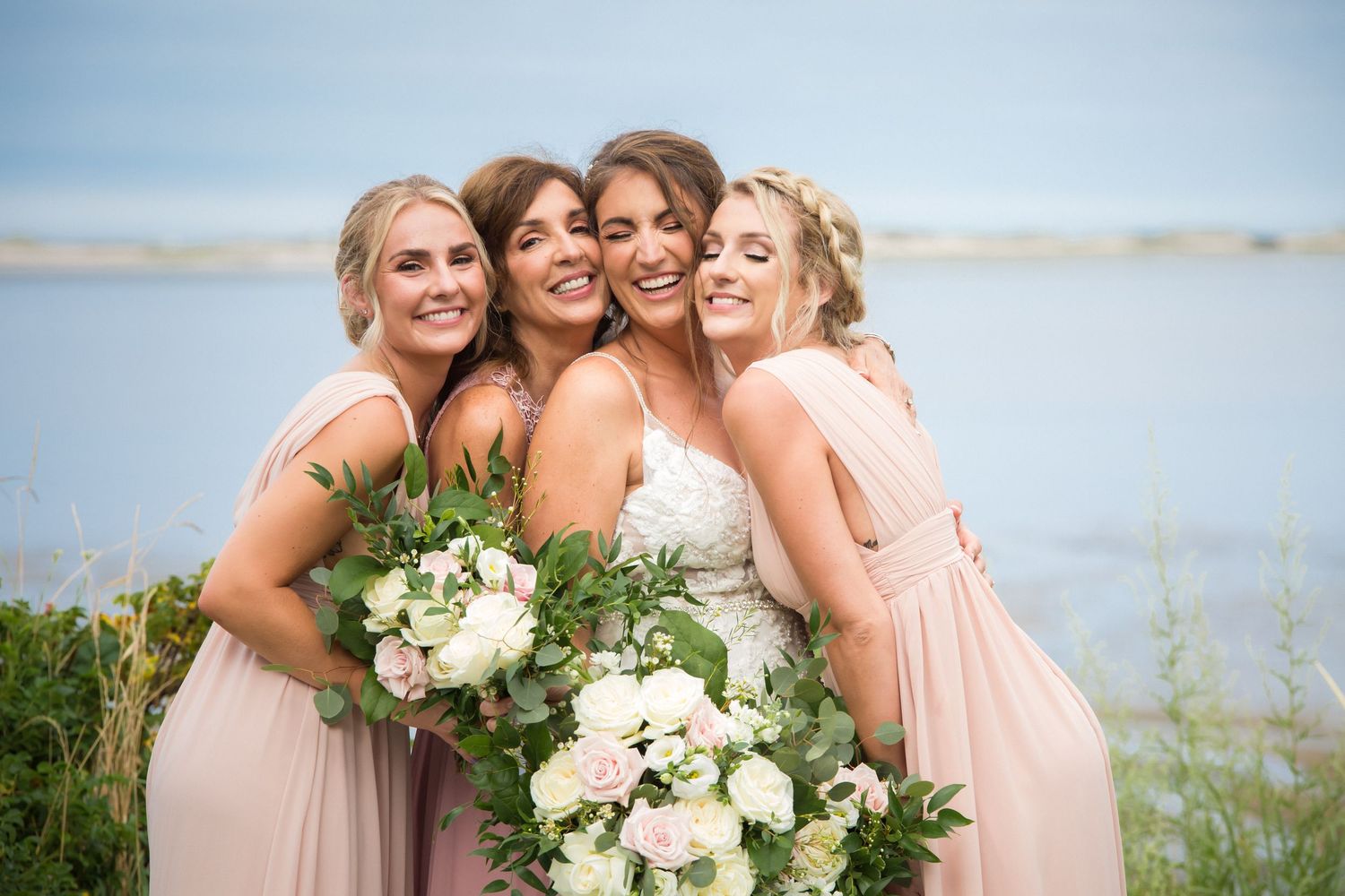 Bride hugs three bridesmaids in blush dresses holding white floral bouquets near waterfront, East Bay Grille, Plymouth, MA