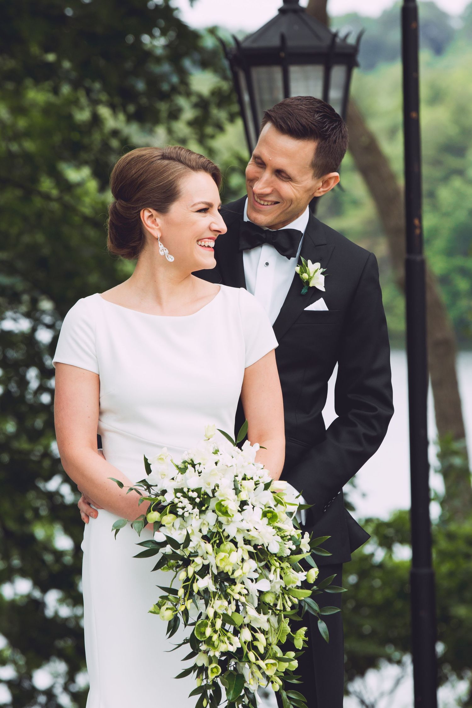 A smiling bride holding a lush white floral bouquet stands with her tuxedoed groom near a garden lamppost, Wellesley College, Wellesley, MA