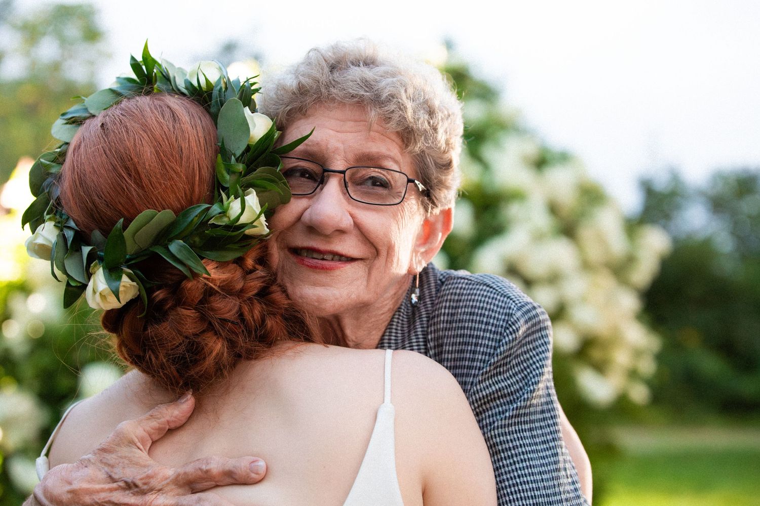 Elderly woman with glasses warmly embraces redhead bride wearing floral flower crown outdoors, Xenia Helix Photography, Boston, MA