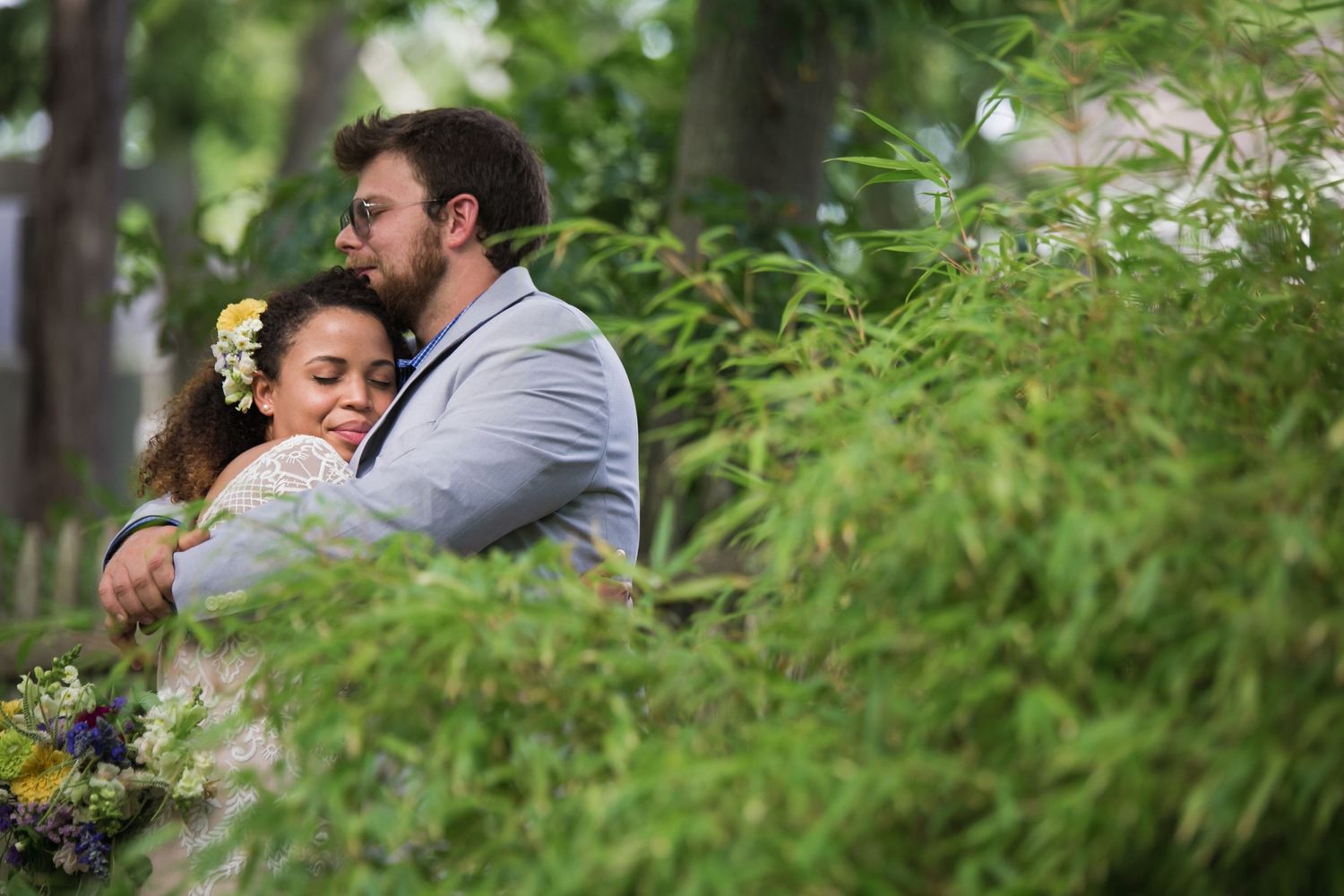Couple embracing among lush green foliage, bride wearing floral hair accessories on their wedding day, Xenia Helix Photography, Boston, MA