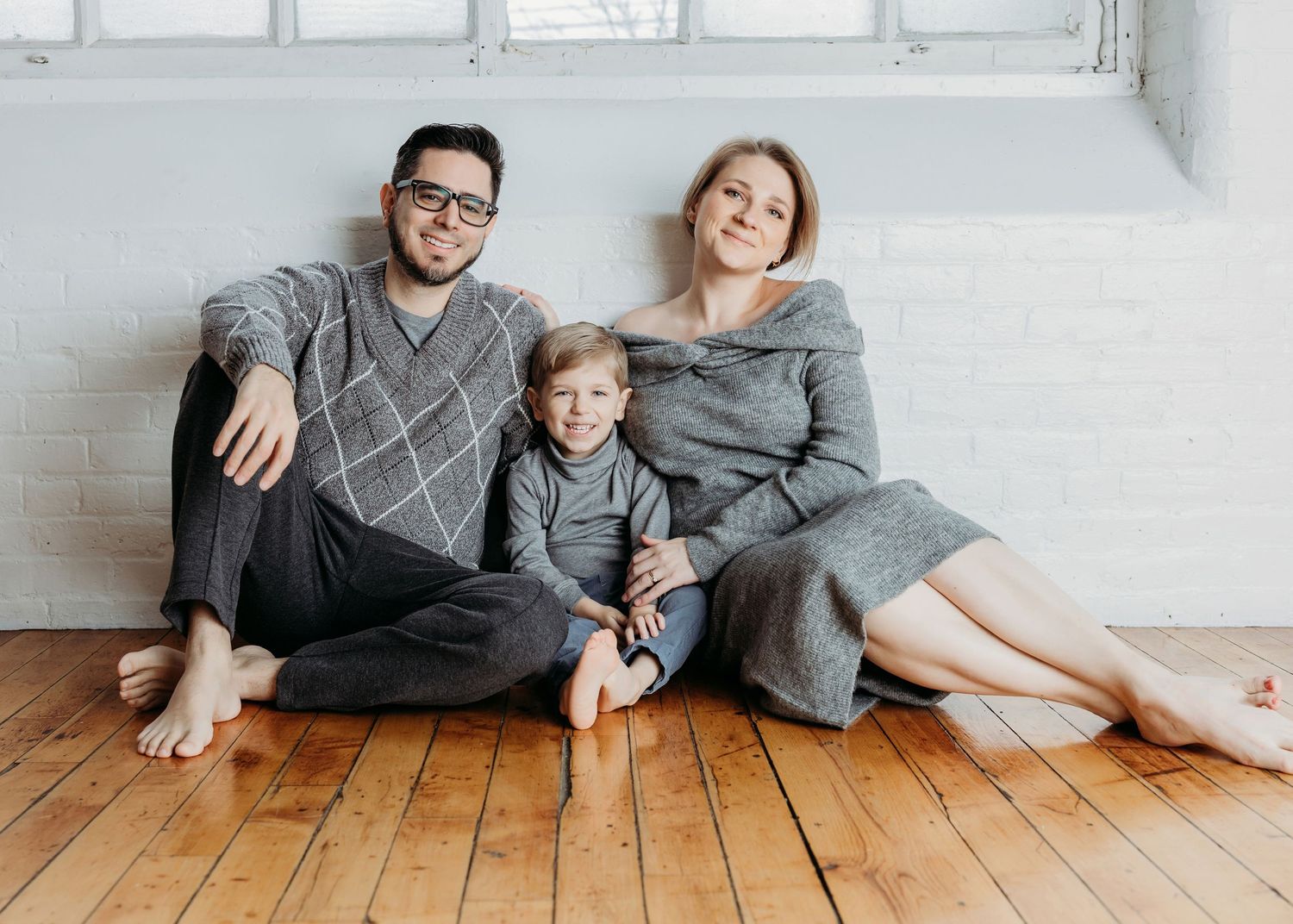 Happy family of three in matching grey outfits sit together on a wooden floor.