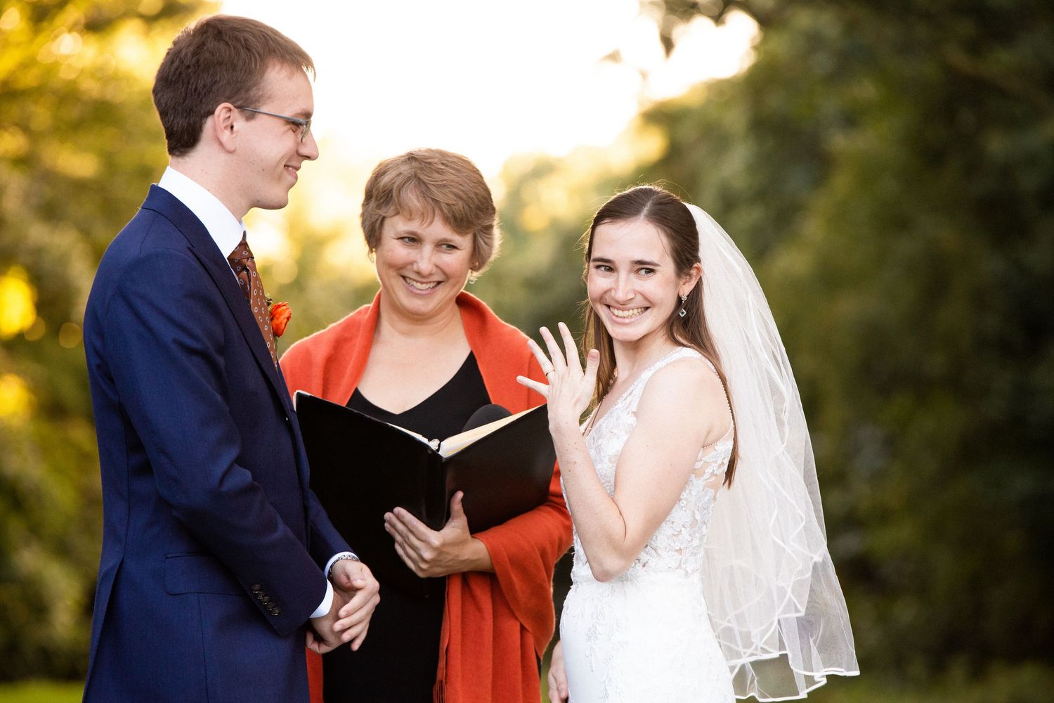 Bride shows off wedding ring while officiant reads ceremony vows outdoors in golden light, The Bradley Estate, Canton, MA