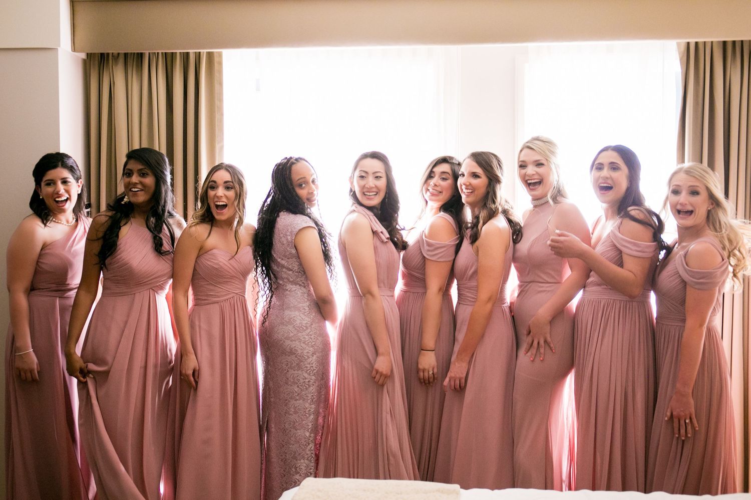 Nine bridesmaids in dusty rose gowns laugh and pose together in a bright hotel room, Mandarin Oriental Boston, Boston, MA