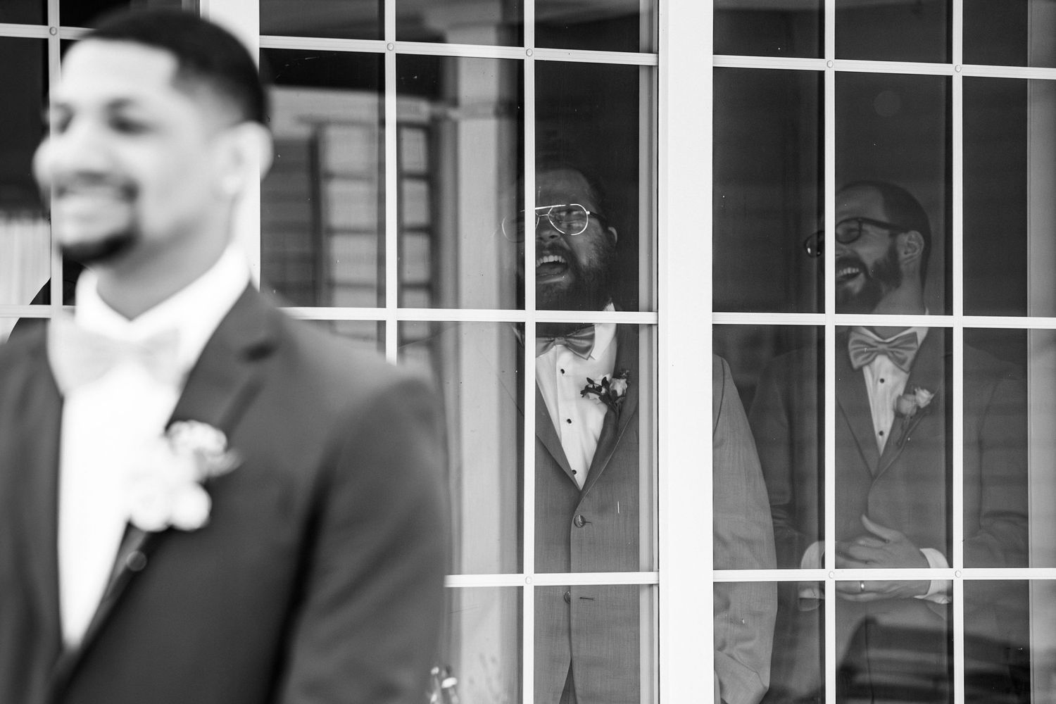 Groomsmen peer through glass doors smiling at groom in black and white wedding photo, Groveland Fairways, Groveland, MA