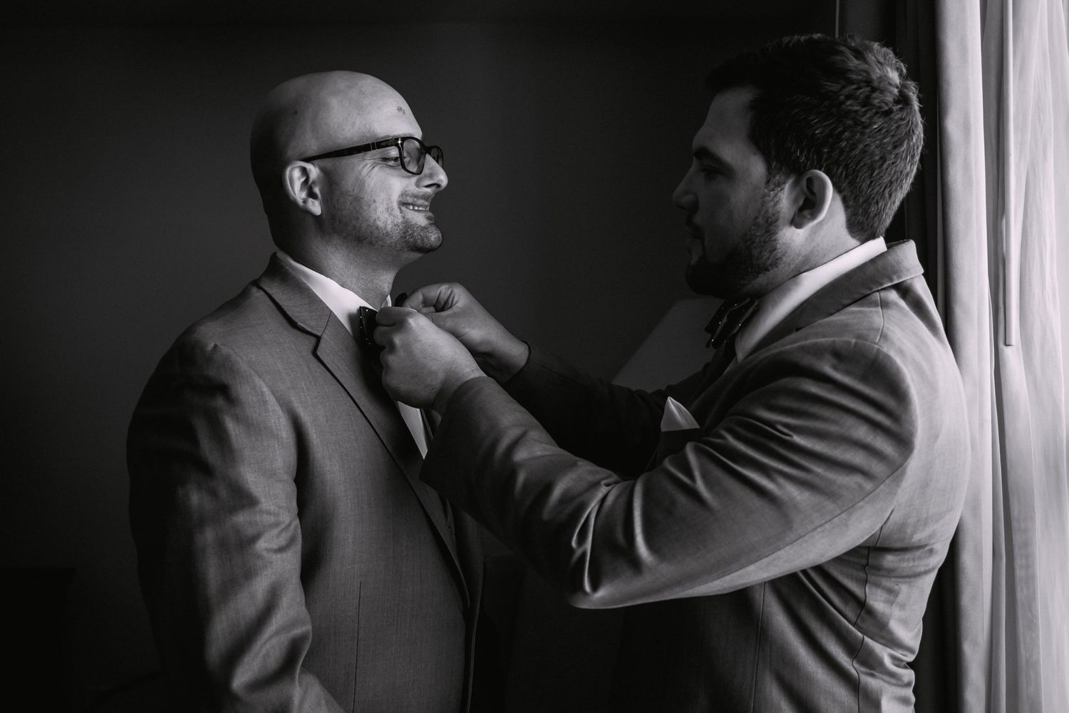 Black and white photo of groomsman adjusting groom's bow tie near window before wedding ceremony, Xenia Helix Photography, Boston, MA