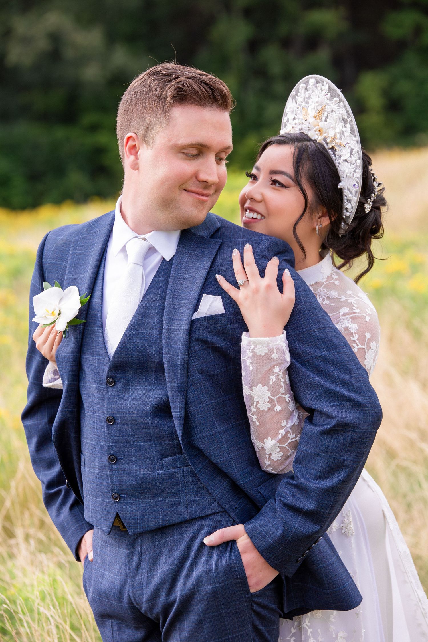 Smiling bride in a lace Vietnamese gown embraces groom in a blue suit in a sunny field, Harrington Farm, Princeton, MA