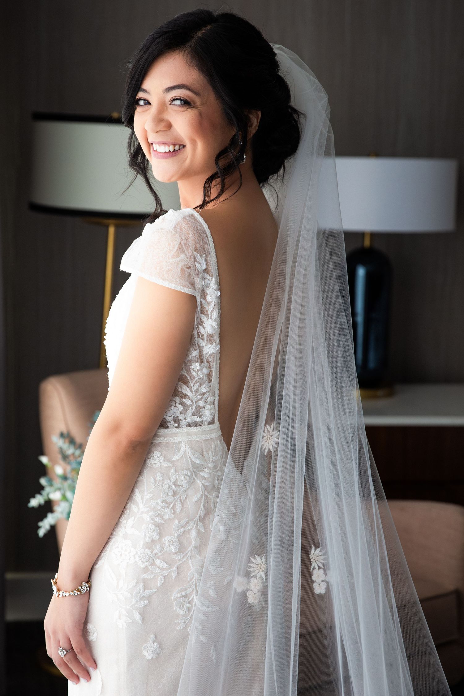 Radiant bride in backless lace wedding dress with 3D floral veil smiling over her shoulder, The Colonnade Hotel, Boston, MA