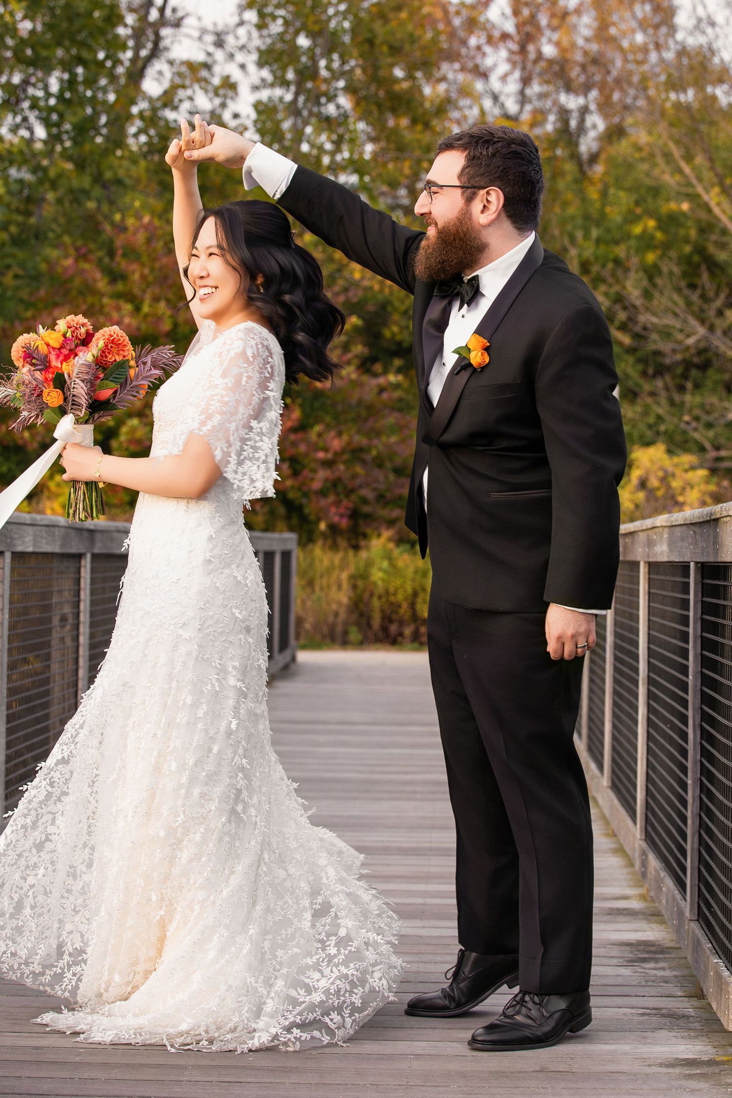 Groom twirls bride in white lace gown on wooden bridge surrounded by vibrant autumn foliage, Alewife Brook Reservation, Cambridge, MA