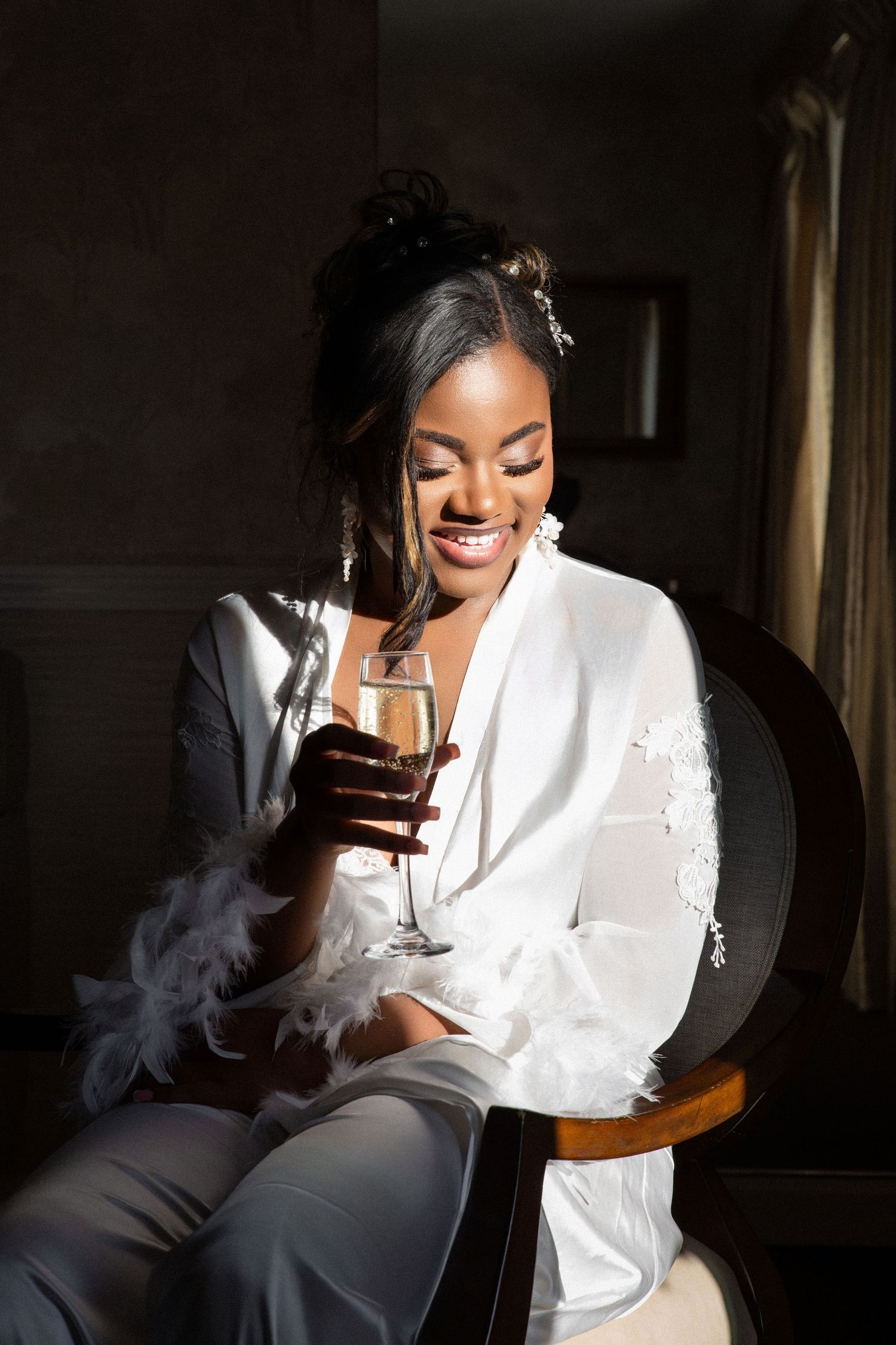 Smiling Black bride in white feather robe holding champagne glass in sunlit room, Independence Harbor, Assonet, MA