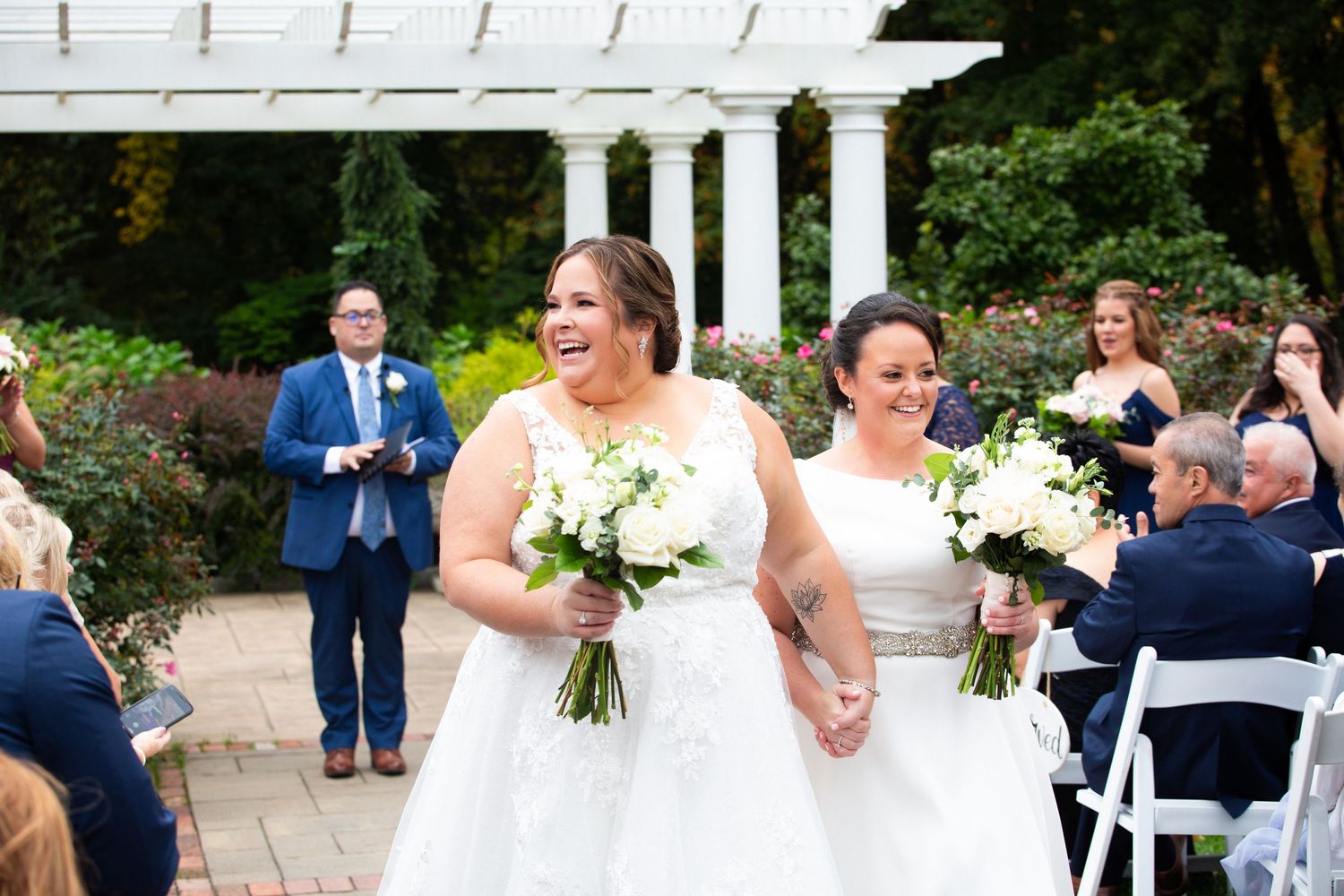 Two brides in white gowns holding bouquets walk hand in hand after their garden ceremony, Coachmen's Lodge, Bellingham, MA