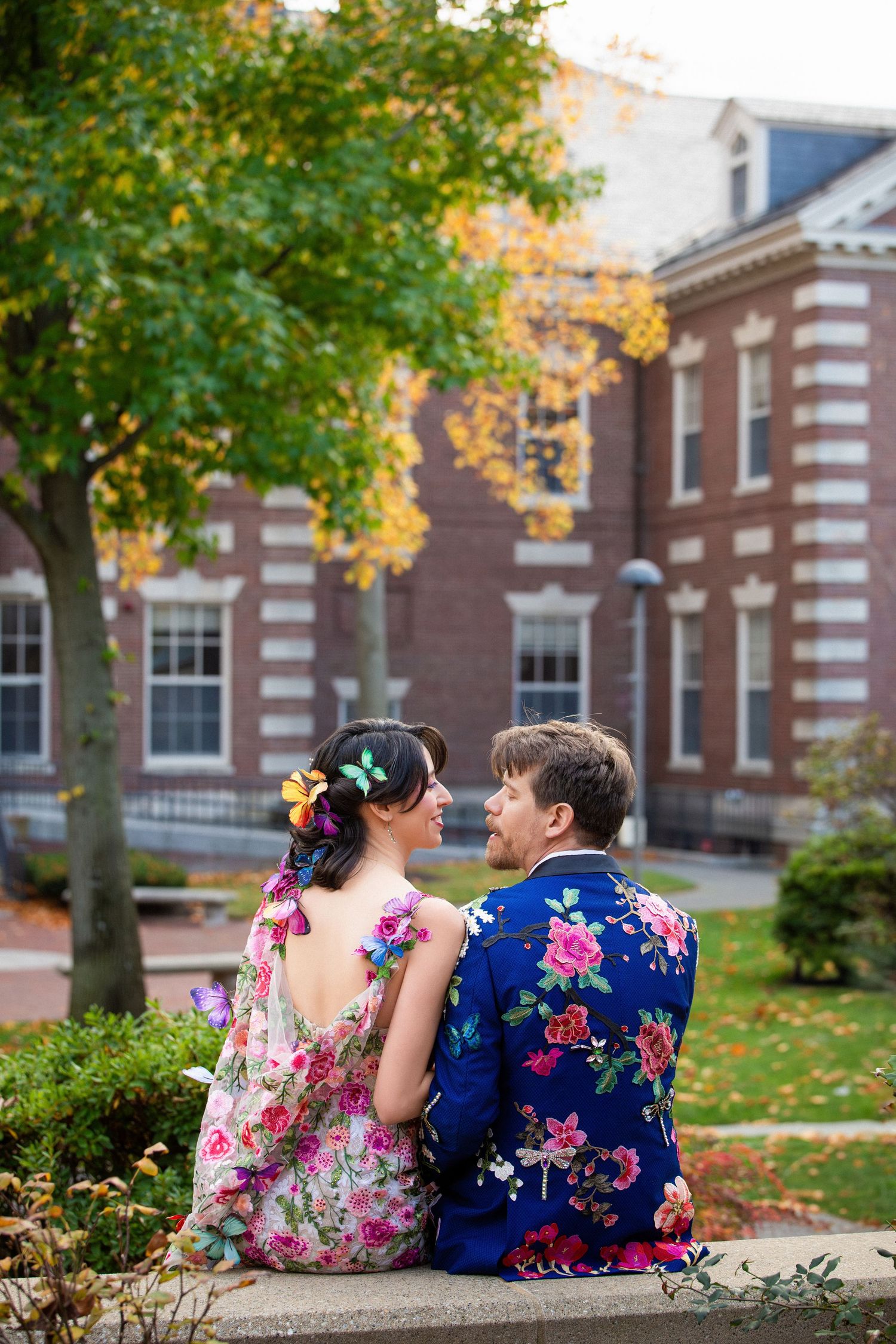 Bride & groom in matching floral outfits sit together in autumn garden setting outside a brick building, Harvard Faculty Club, Cambridge, MA
