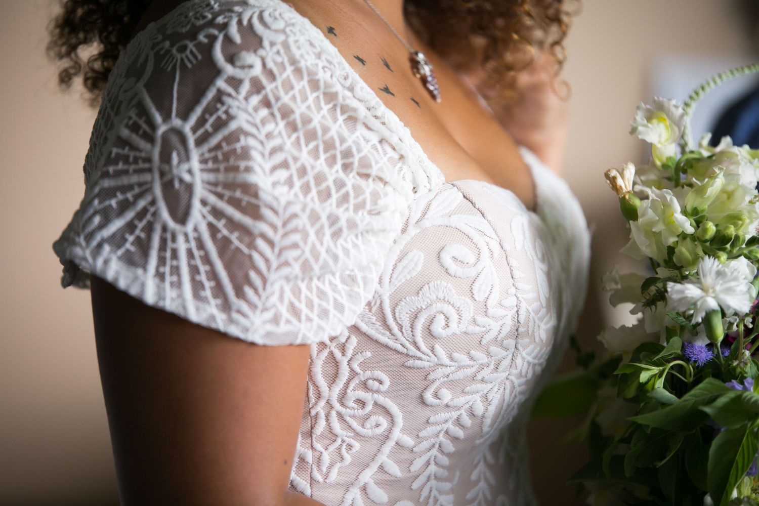 Close-up of a bride with tattoos in an intricate lace cap sleeve wedding gown holding a floral bouquet, Xenia Helix Photography, Boston, MA