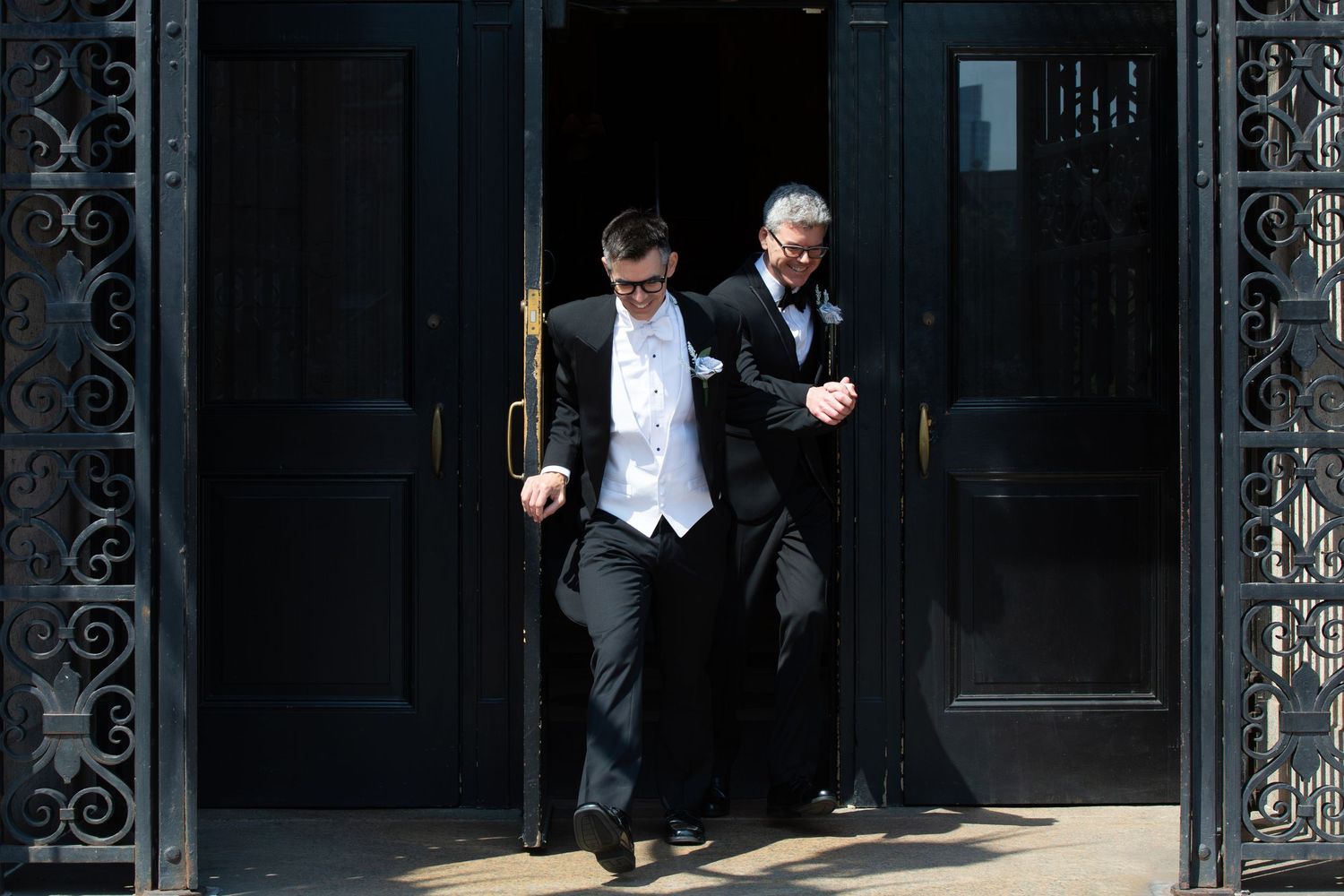 Two grooms in formal tuxedos emerge through grand ornate black doors on their wedding day, Boston Public Library, Boston, MA