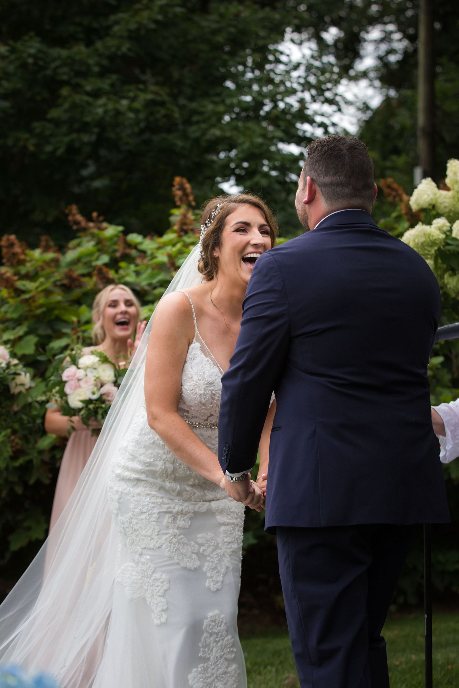 Joyful bride in lace gown laughing while holding grooms hands during outdoor garden ceremony, Brewster Gardens, Plymouth, MA