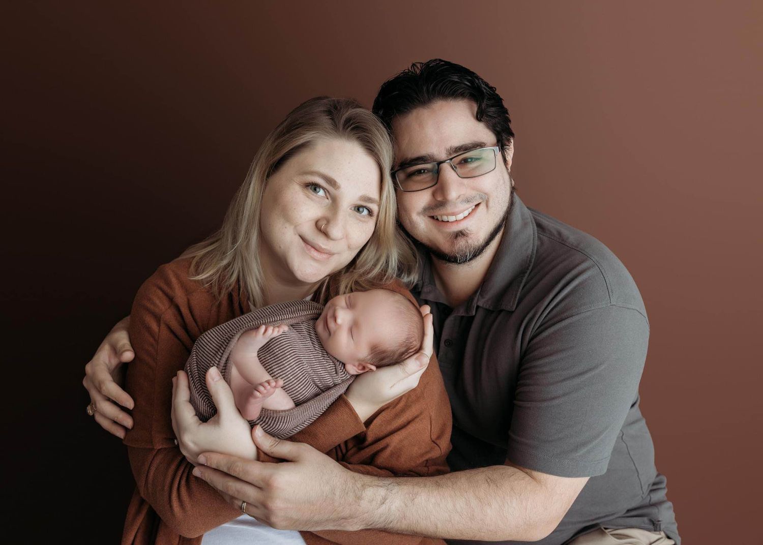 Xenia and Cesar smile while cradling their newborn baby against a warm brown background.