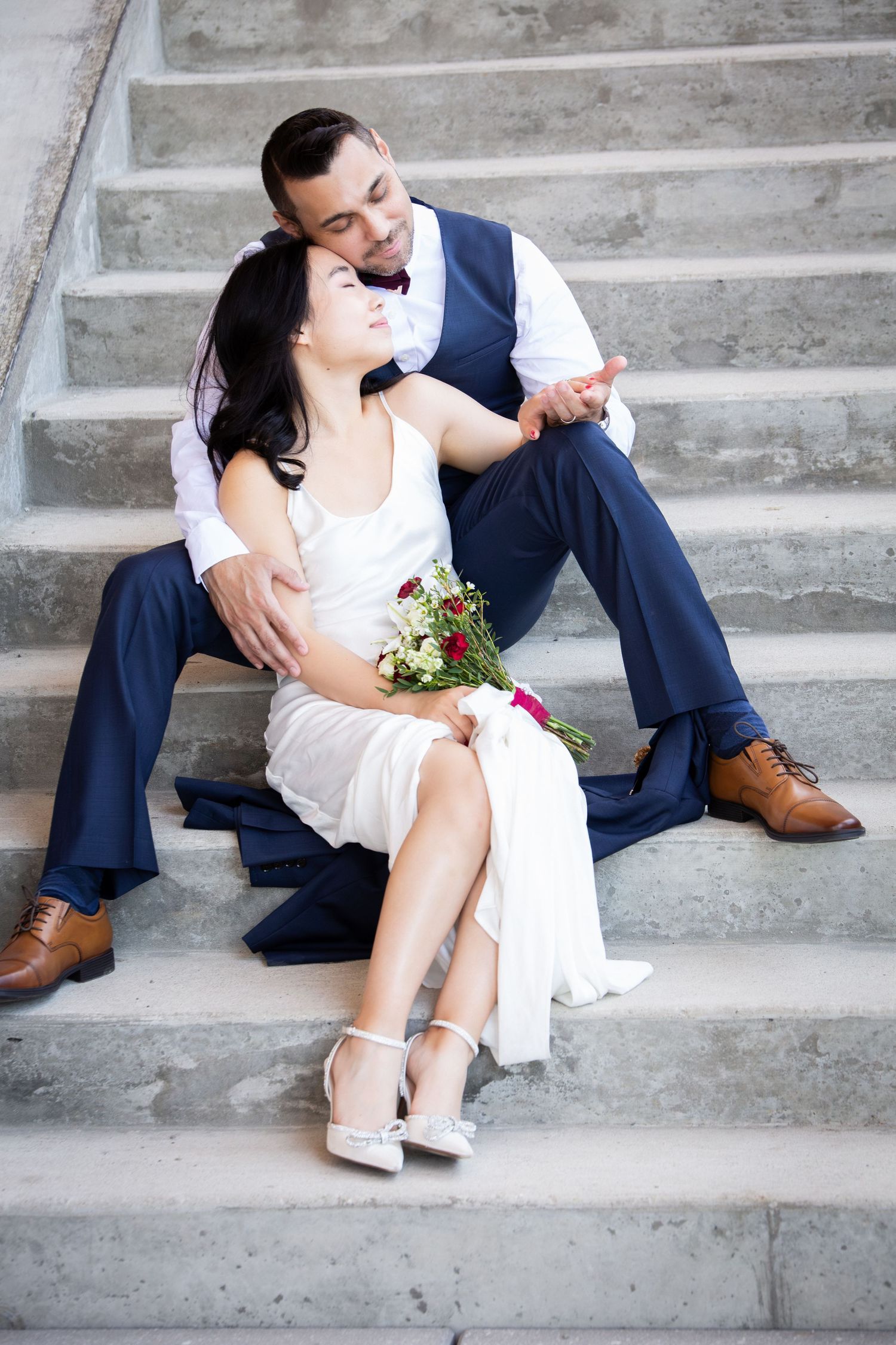 Bride and groom sit together on stone steps, groom embracing bride holding a floral bouquet, Somerville City Hall, Somerville, MA
