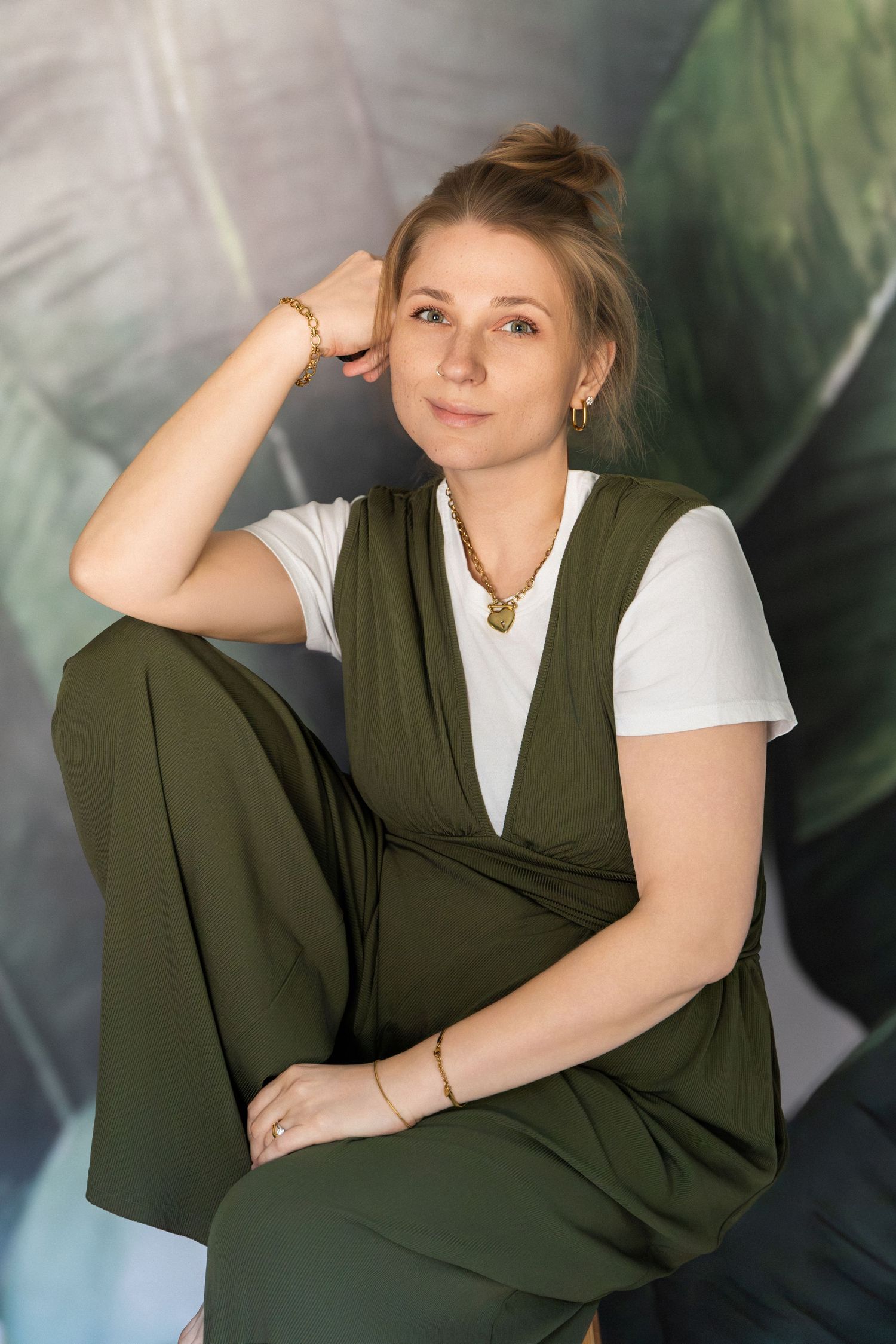 Xenia Helix — Boston-based wedding photographer —  in green jumpsuit with gold jewelry sits relaxed against tropical leaf backdrop.